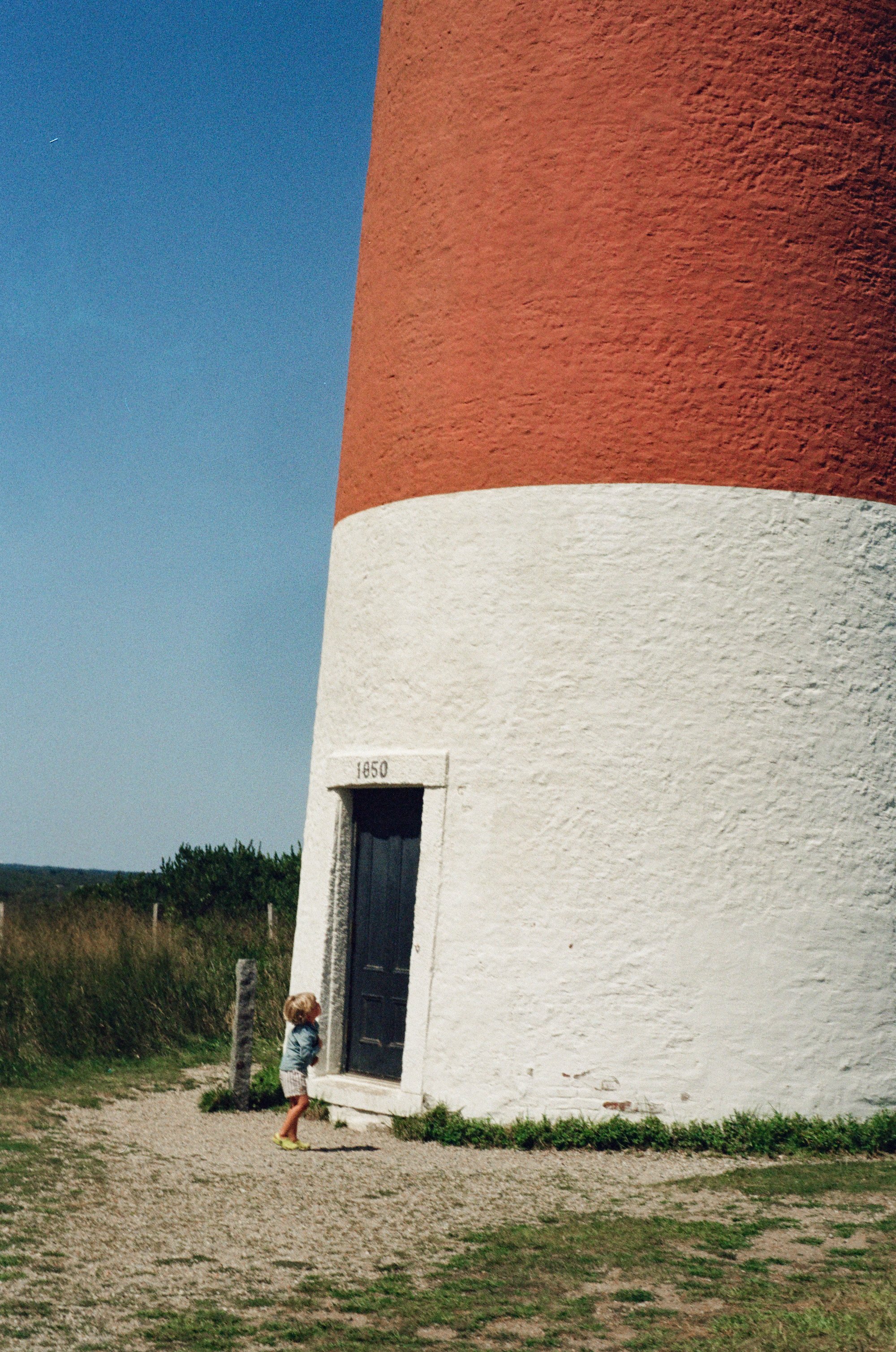 A young girl with blonde hair, wearing a leather jacket, shorts, and yellow shoes, standing outside a large white and red lighthouse, looking up at it.