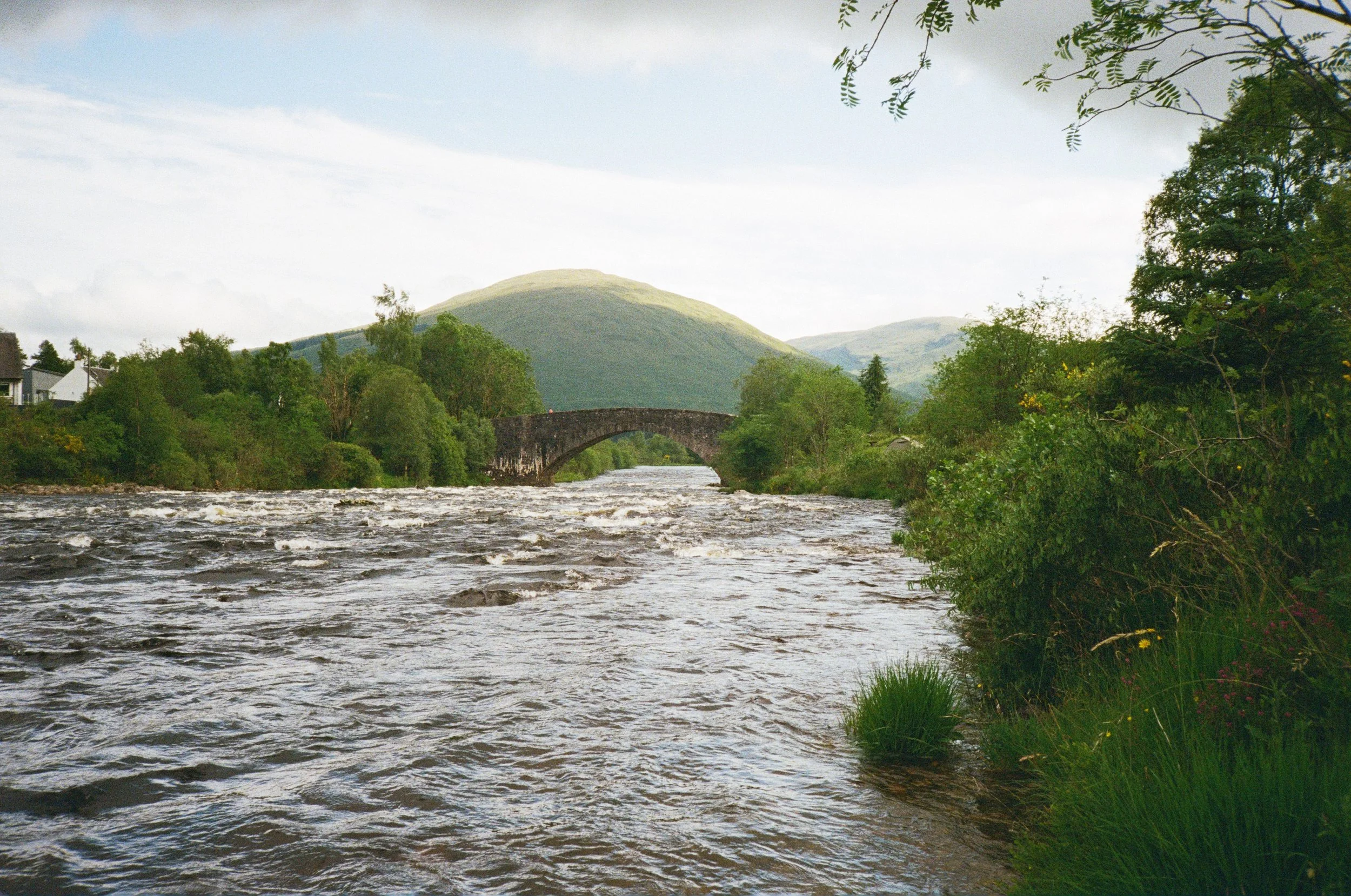 A river flowing through a green landscape with trees, hills, and an old stone bridge under a partially cloudy sky.