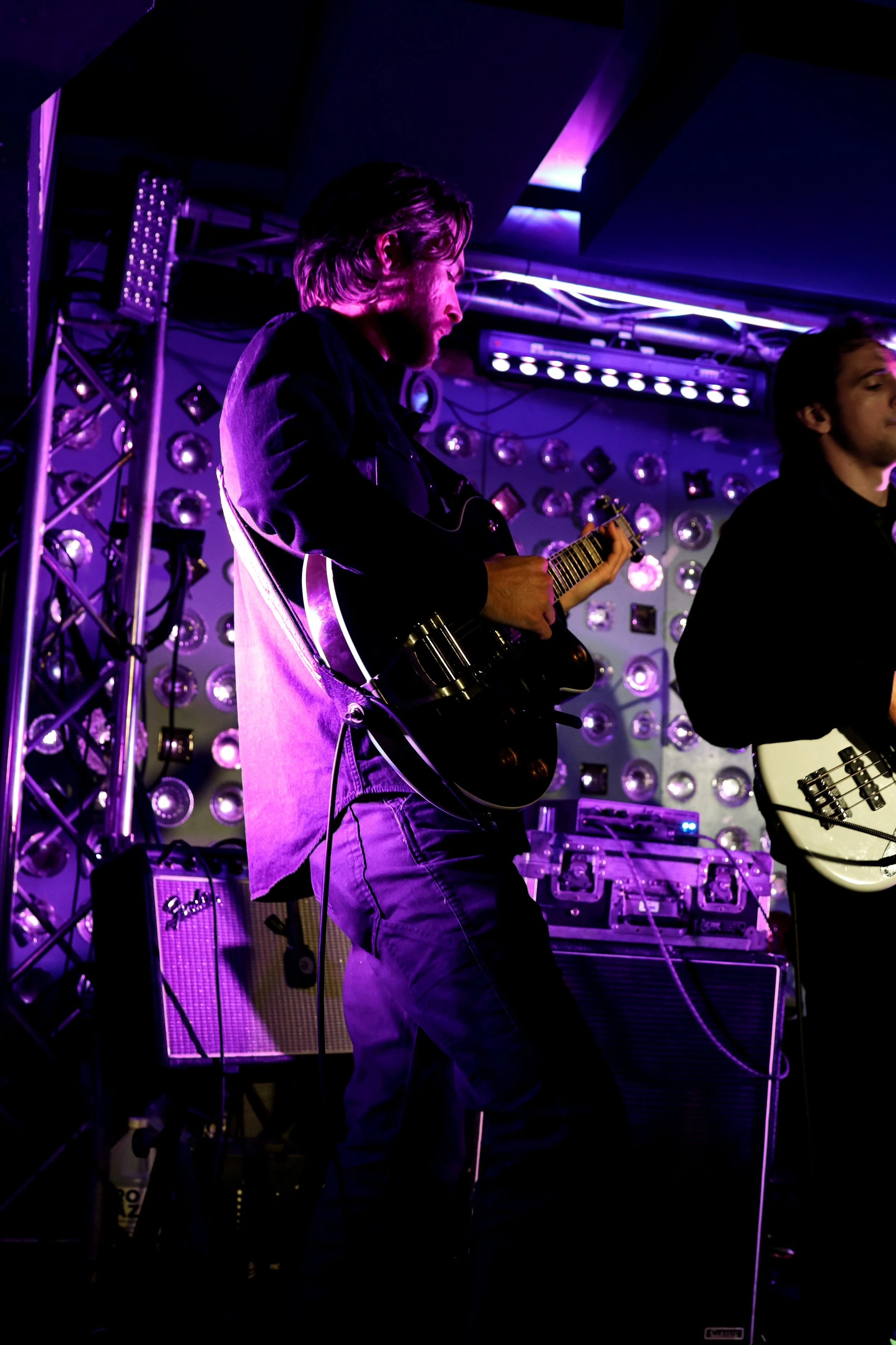 Musicians playing guitars on stage with purple lighting and a patterned wall background.