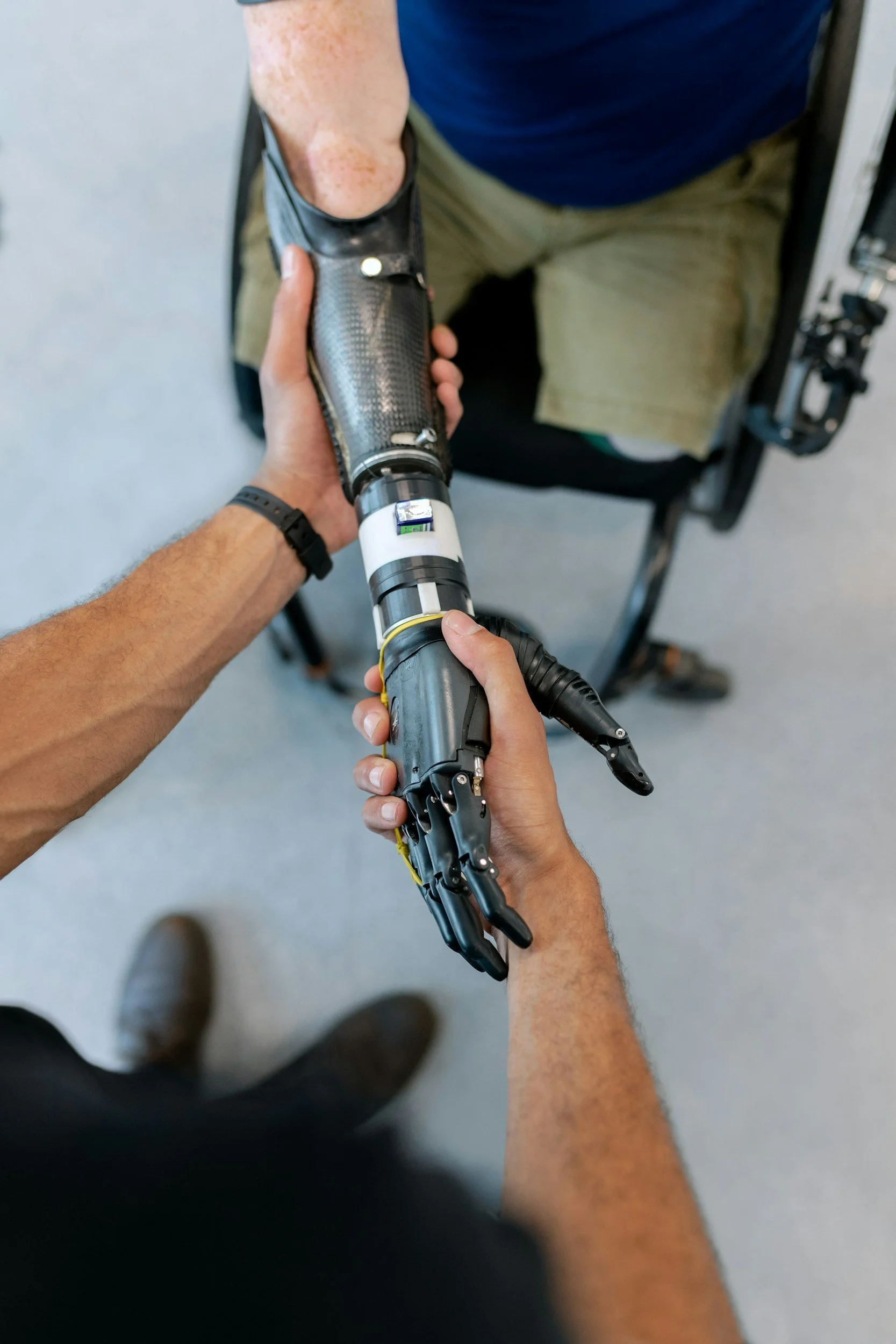 A person with a robotic prosthetic arm held with hands in view, standing on a gray floor with another person in the background wearing khaki shorts and a dark blue shirt.