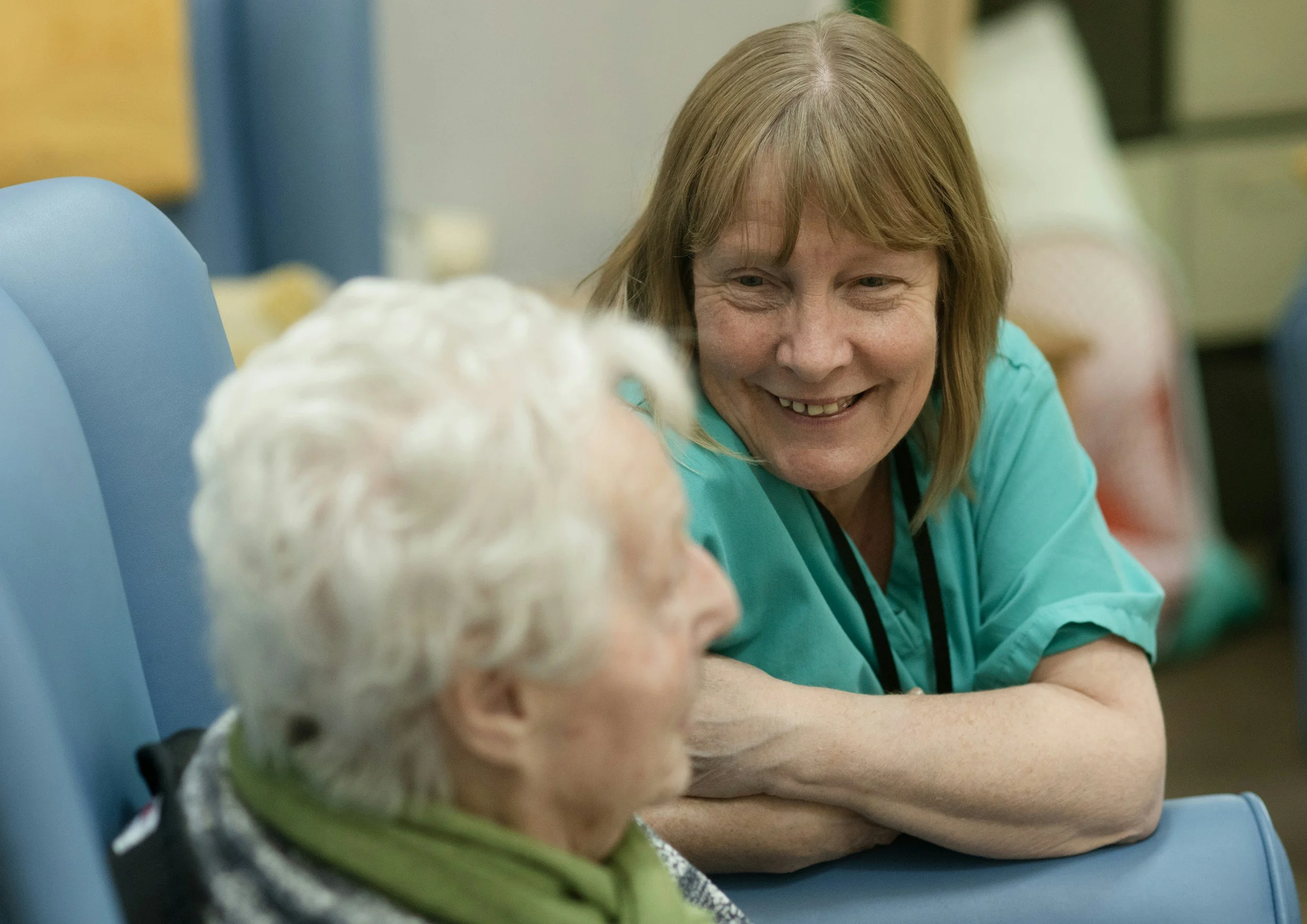 A smiling middle-aged woman with red hair and a turquoise shirt sitting next to an elderly person with white hair, in a colorful setting.