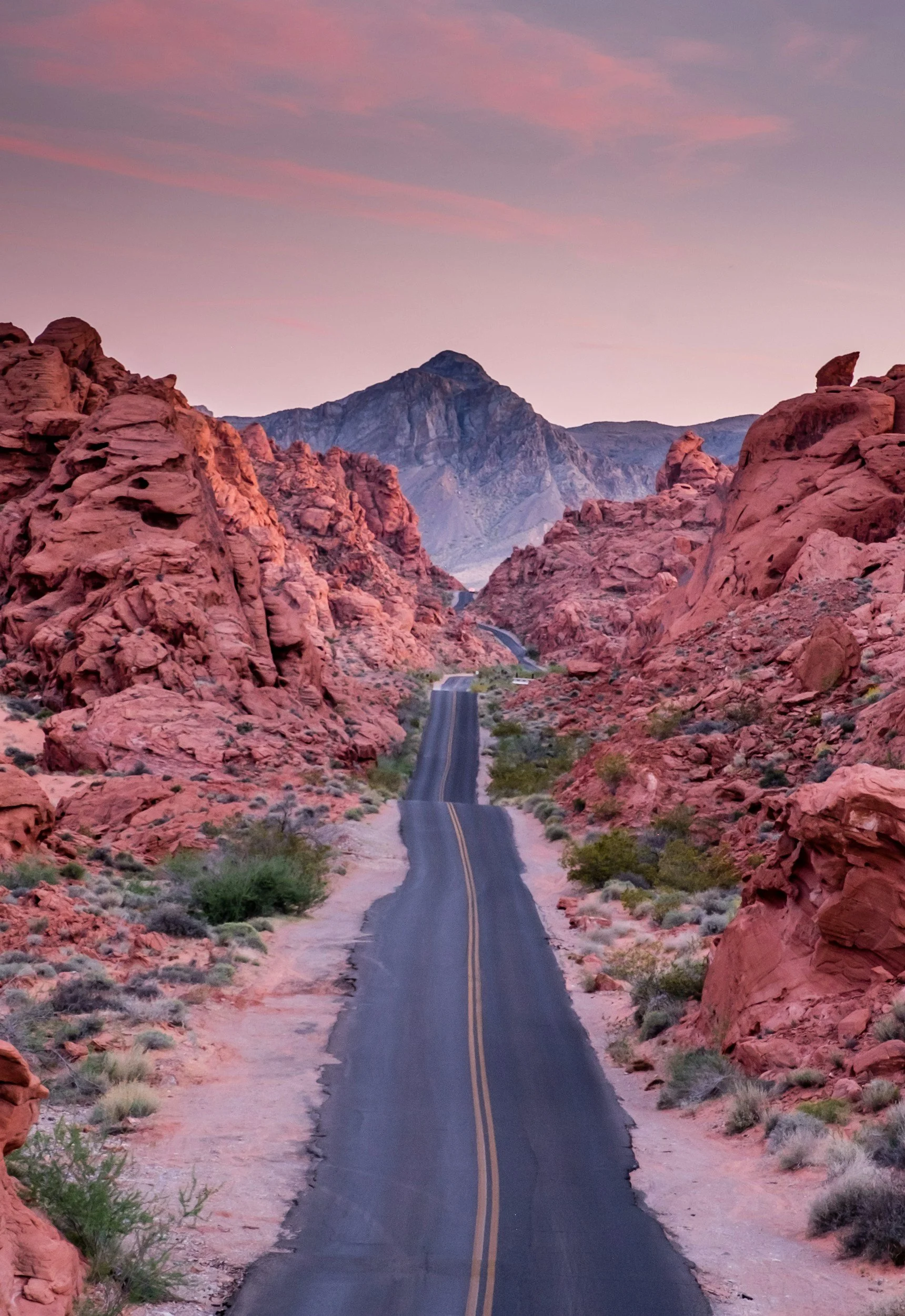 A straight two-lane road running through a canyon with red rock formations, heading towards mountains in the distance under a pink and purple sky.