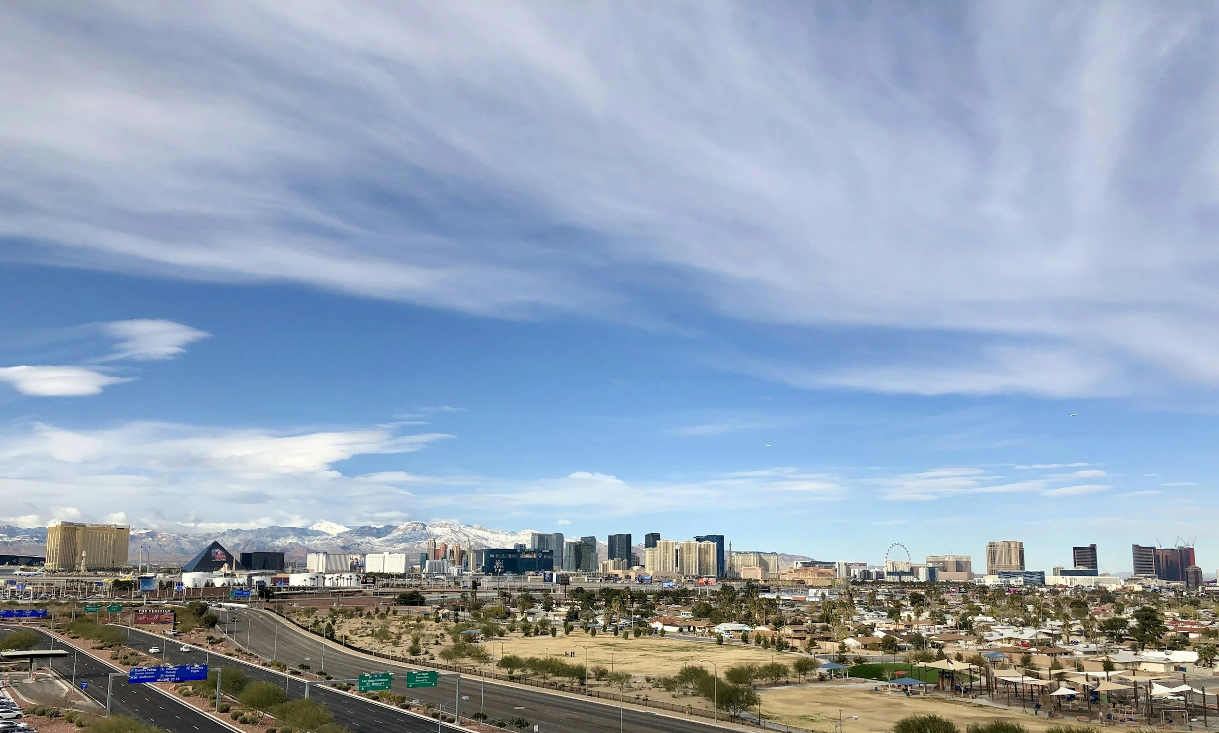 City skyline with high-rise buildings, mountains in the background, blue sky with clouds, and a highway in the foreground.
