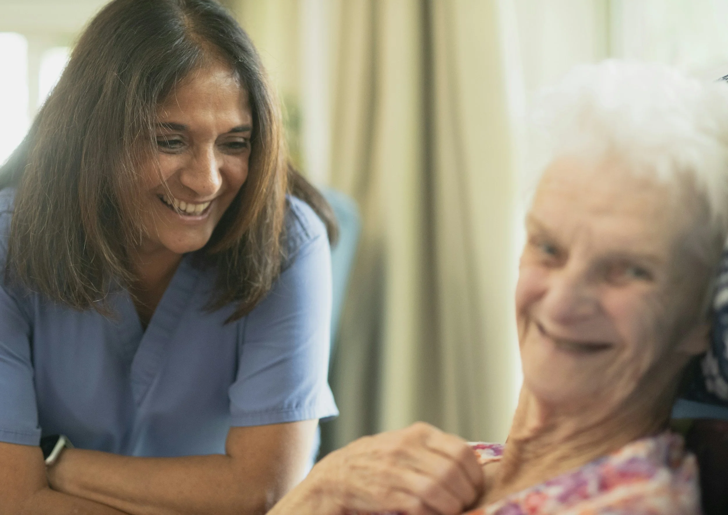 A woman with brown hair smiling, wearing a blue medical scrub, leaning towards an elderly woman with short gray hair, who is also smiling, in a well-lit room with curtains in the background.
