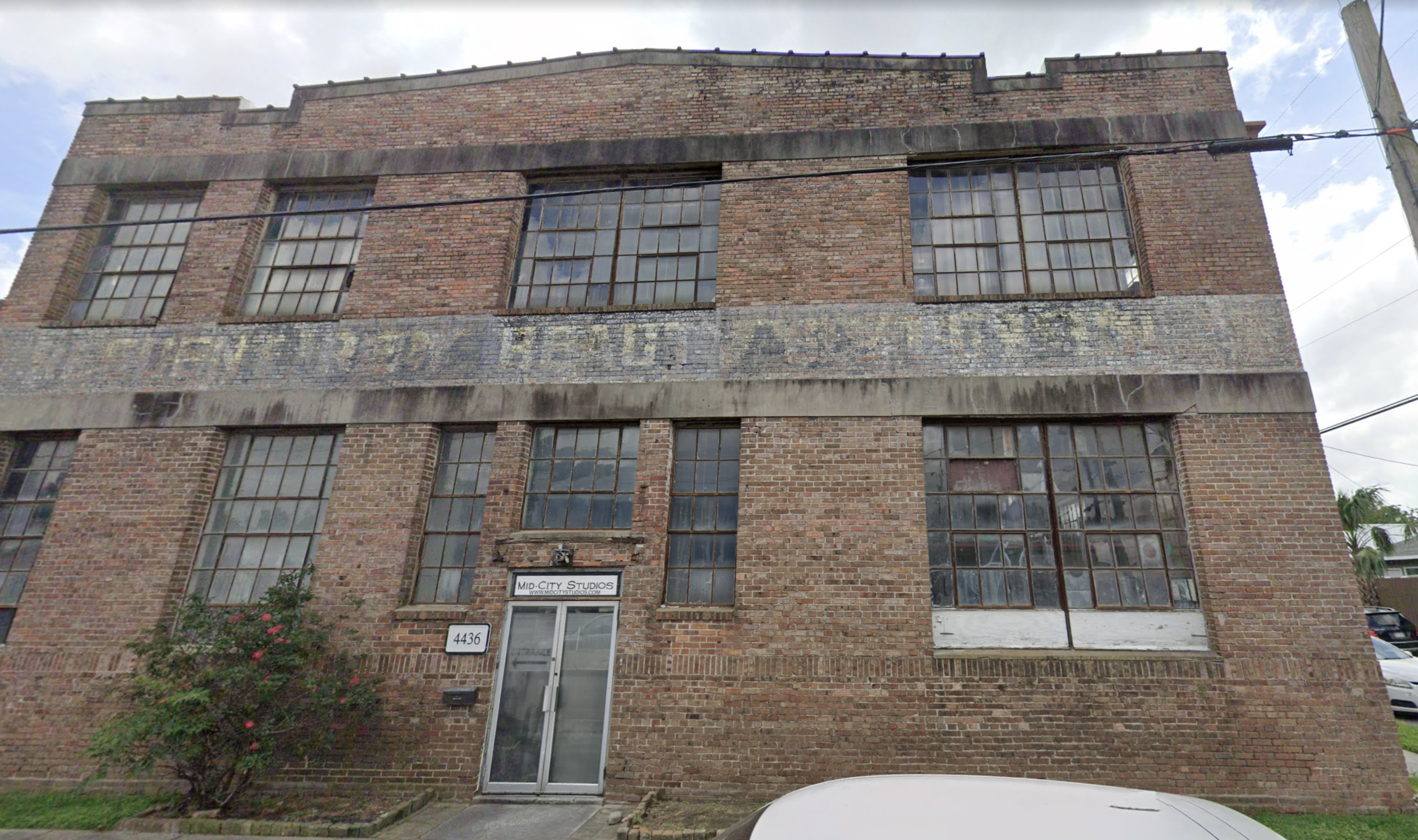 Brick building with large multi-pane windows, a sign that reads 'Mid City Studios,' and a door at the entrance, with a small flower bush and a white car partially visible in front.
