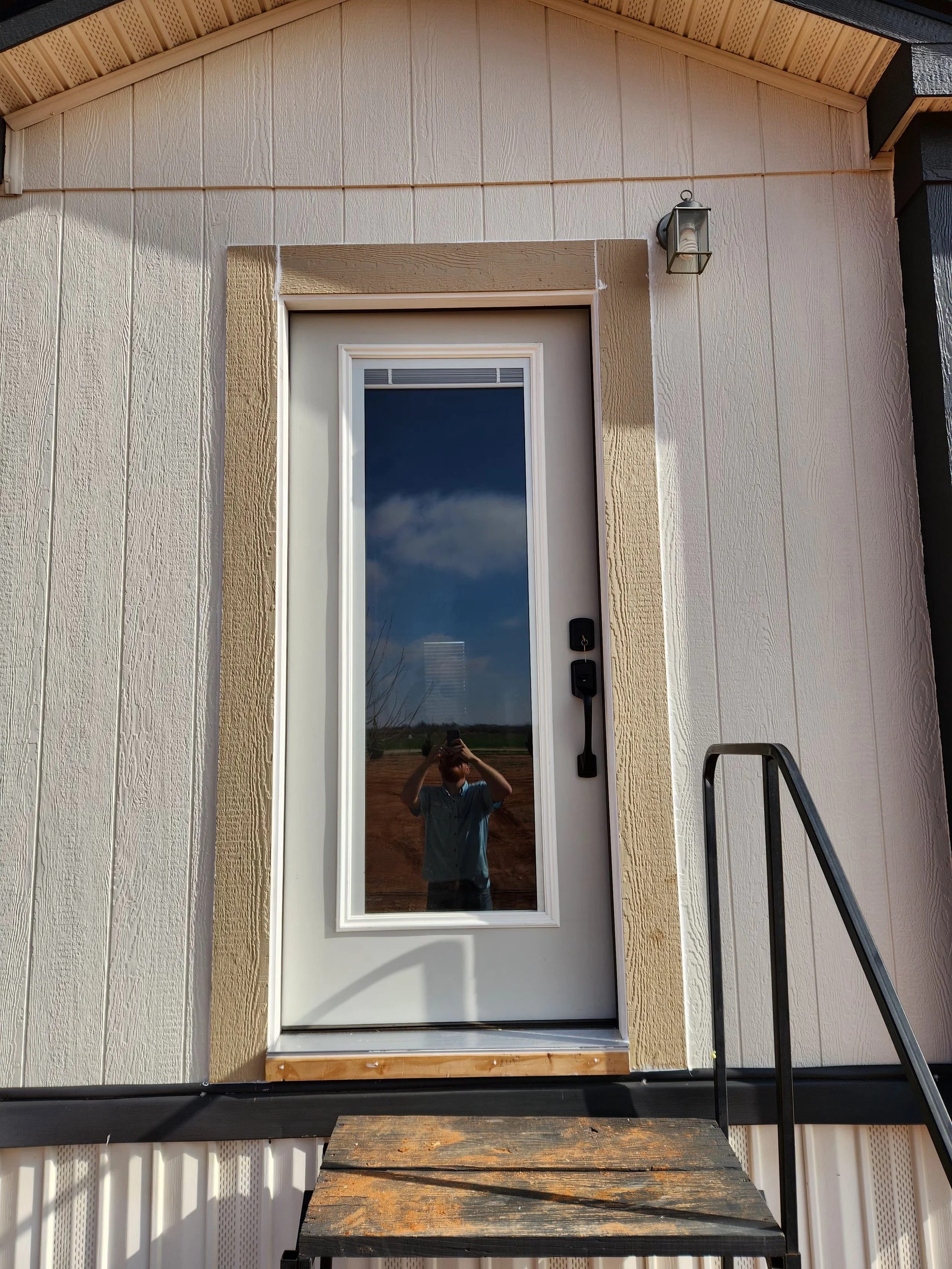 Front door of a house with a glass panel, black handle and lock, surrounded by beige trim, with a wooden step and black metal railing. The reflection in the glass shows a person taking the photo, blue sky, and some clouds.