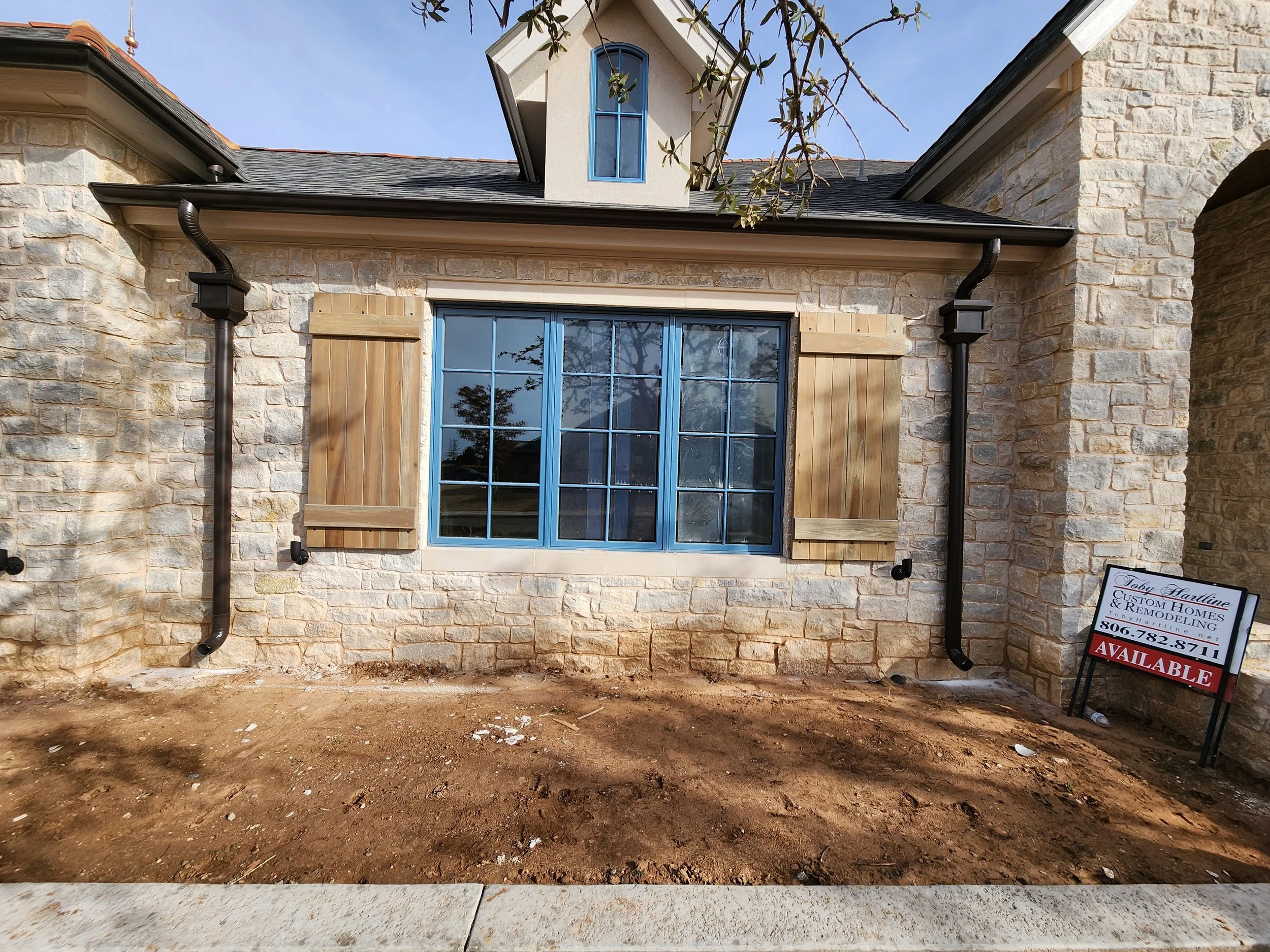 The front of a house under construction with a stone facade, large window with blue frame, and a sign for Toby Hartline & Remodelling company, indicating the house is available. The house has a dormer window, black gutter pipes, and a dirt yard in front.