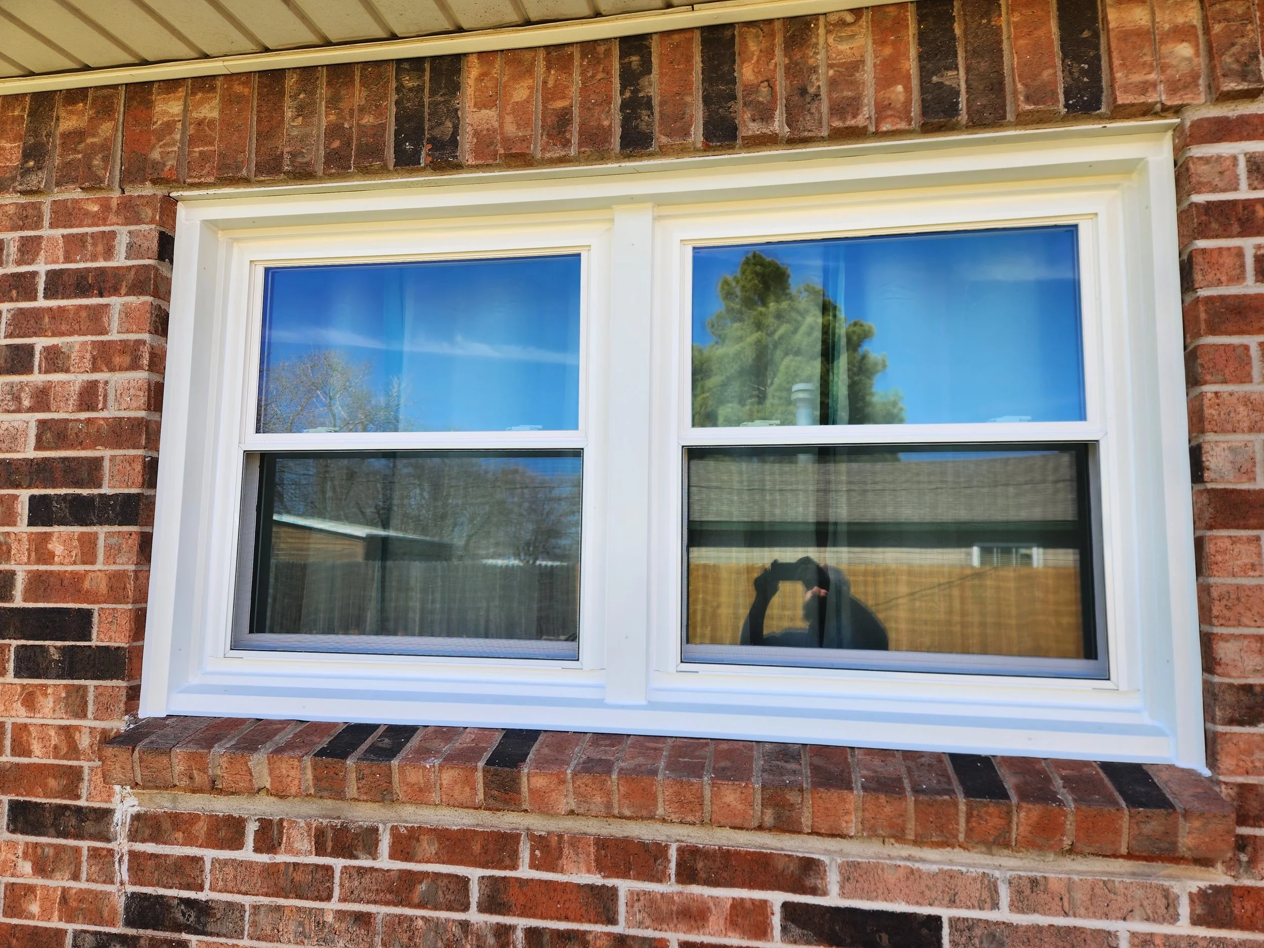 Photo of a double-hung window with a white frame set in a brick wall. The reflection in the glass shows a blue sky, trees, and a person taking the photo.