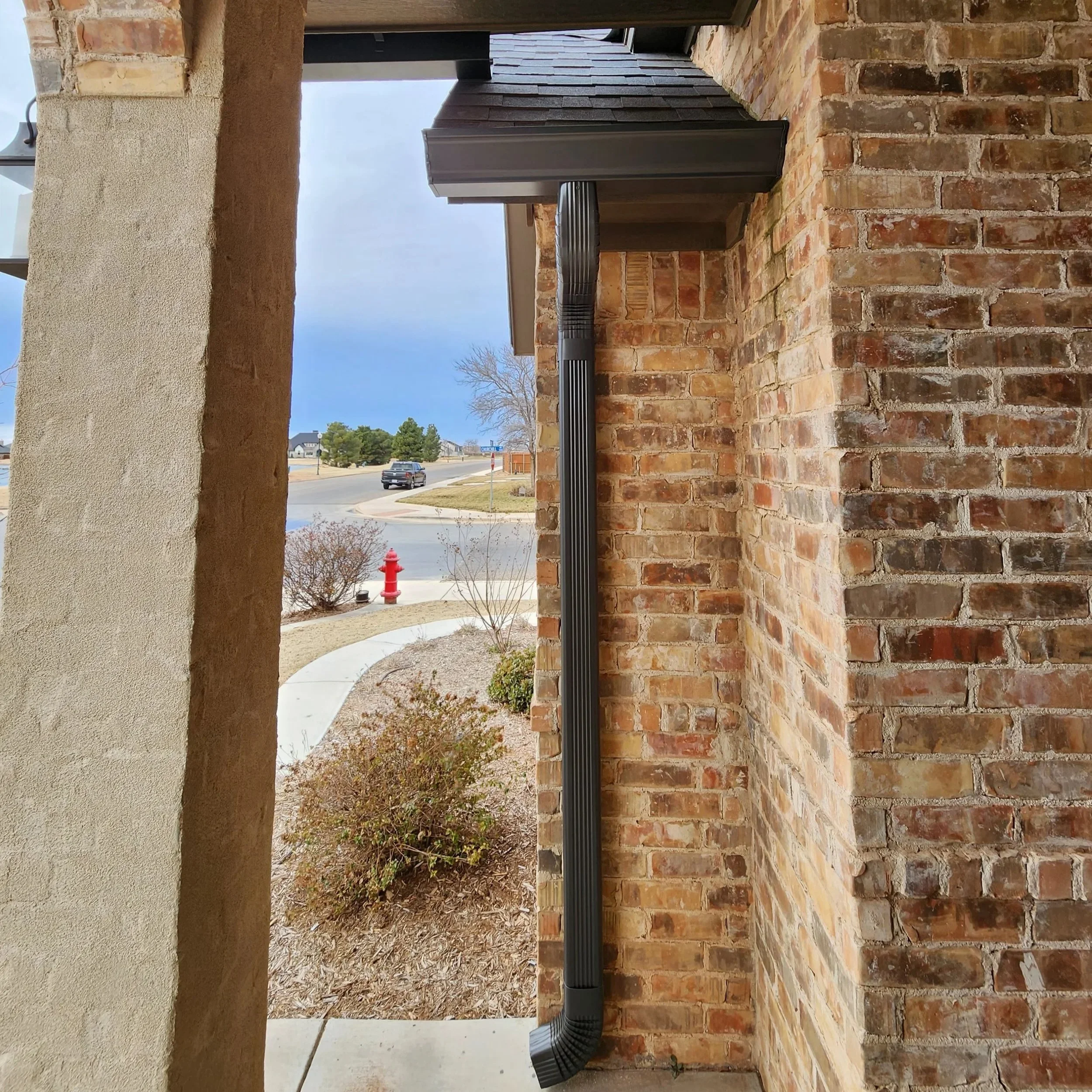 View of a brick porch with a black drainage pipe and a sidewalk with a parked car, street signs, and trees in the background under a partly cloudy sky.