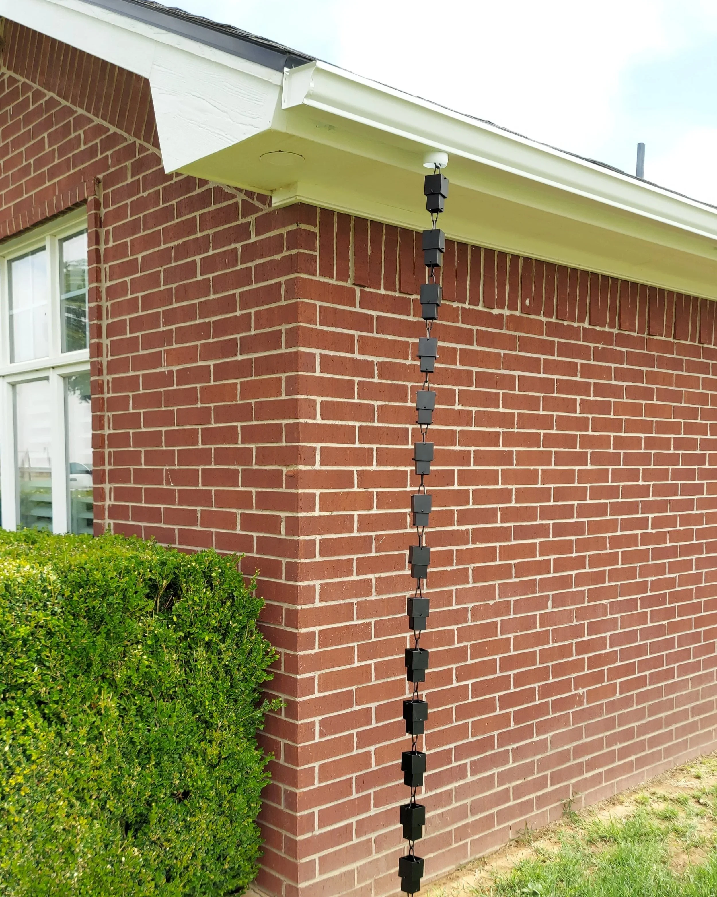 Exterior corner of a brick house with a window, white trim, and a pitched roof. A long string of small, black, cube-shaped objects hangs vertically from the eaves.