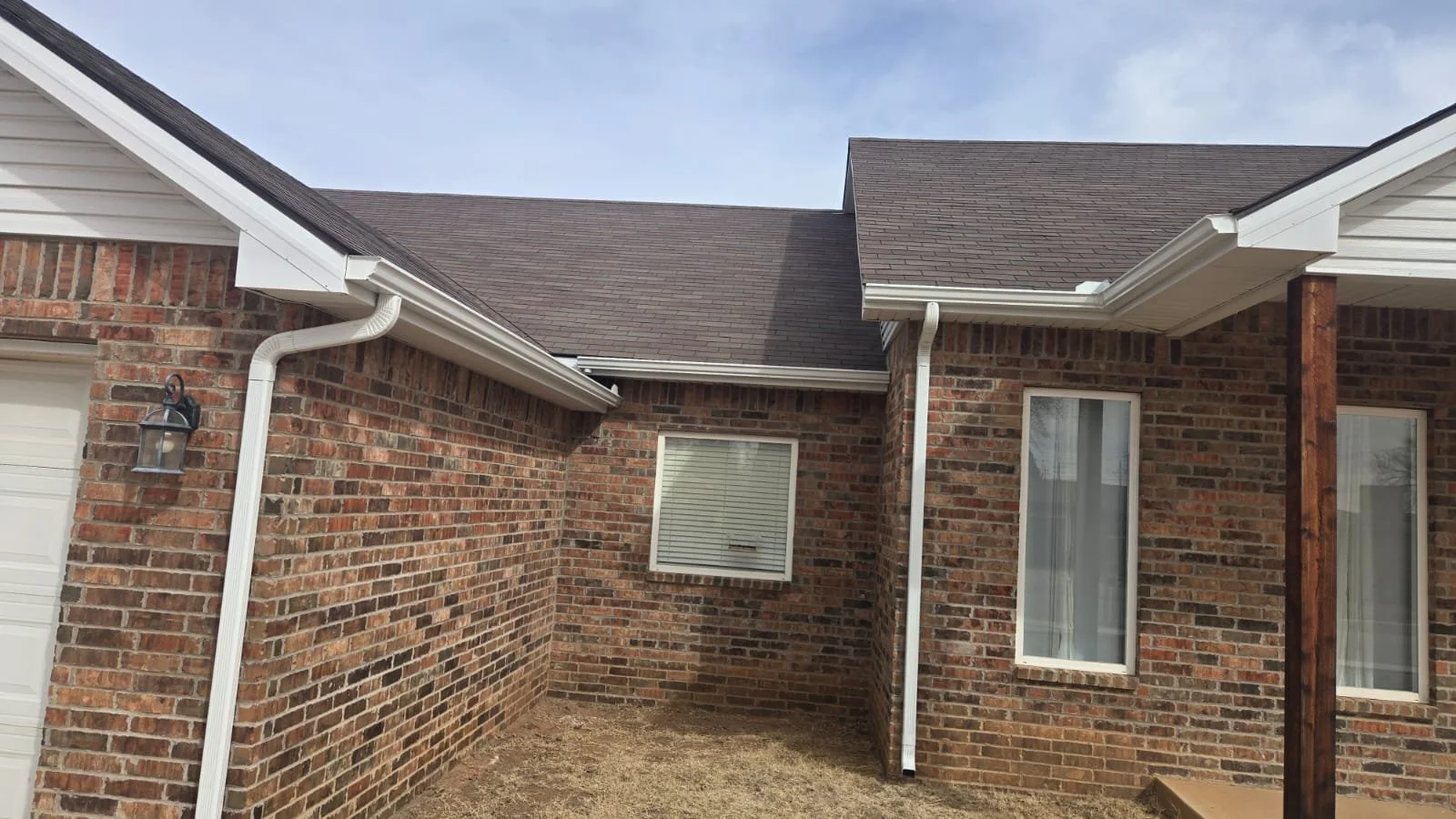 Side view of a brick house with windows, white gutters, and a brown shingled roof, with a small patch of lawn and a lamp mounted on the exterior wall.