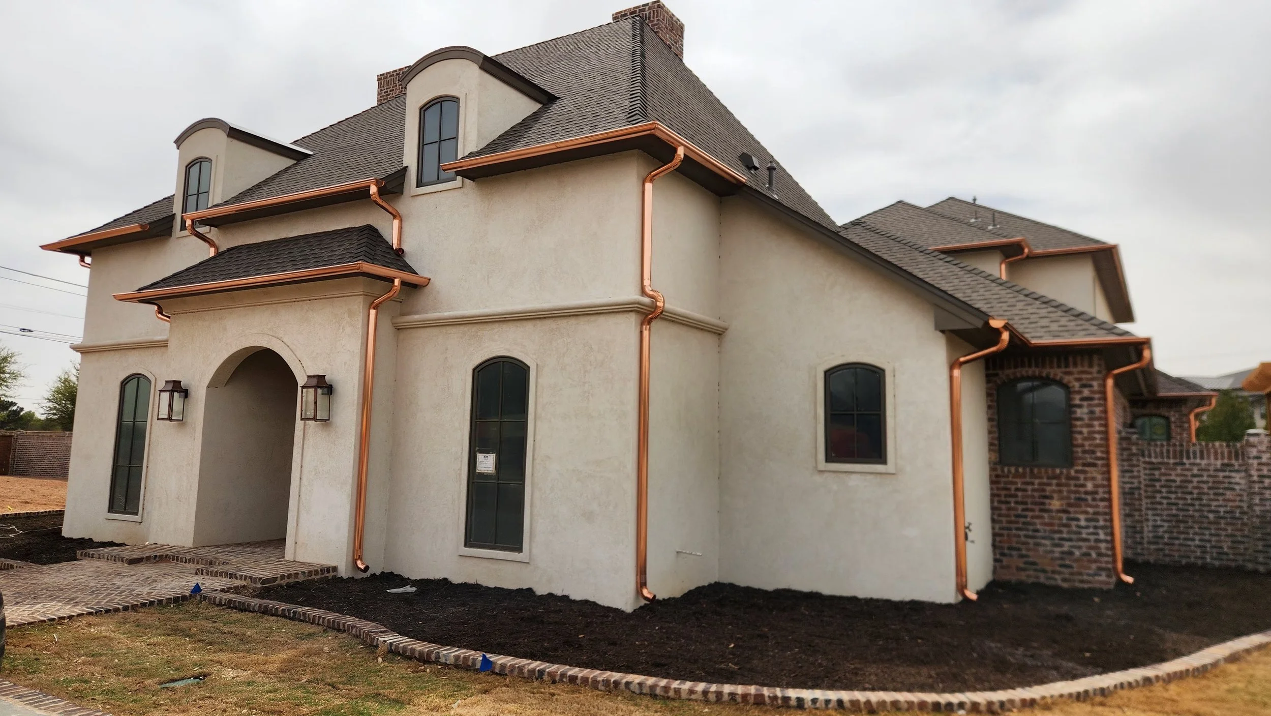 A newly constructed house with a stucco exterior, arched windows, and brick accents. Copper gutters and downspouts are installed along the roofline. The front yard is landscaped with a brick border, and there is a paved walkway leading to the entrance.