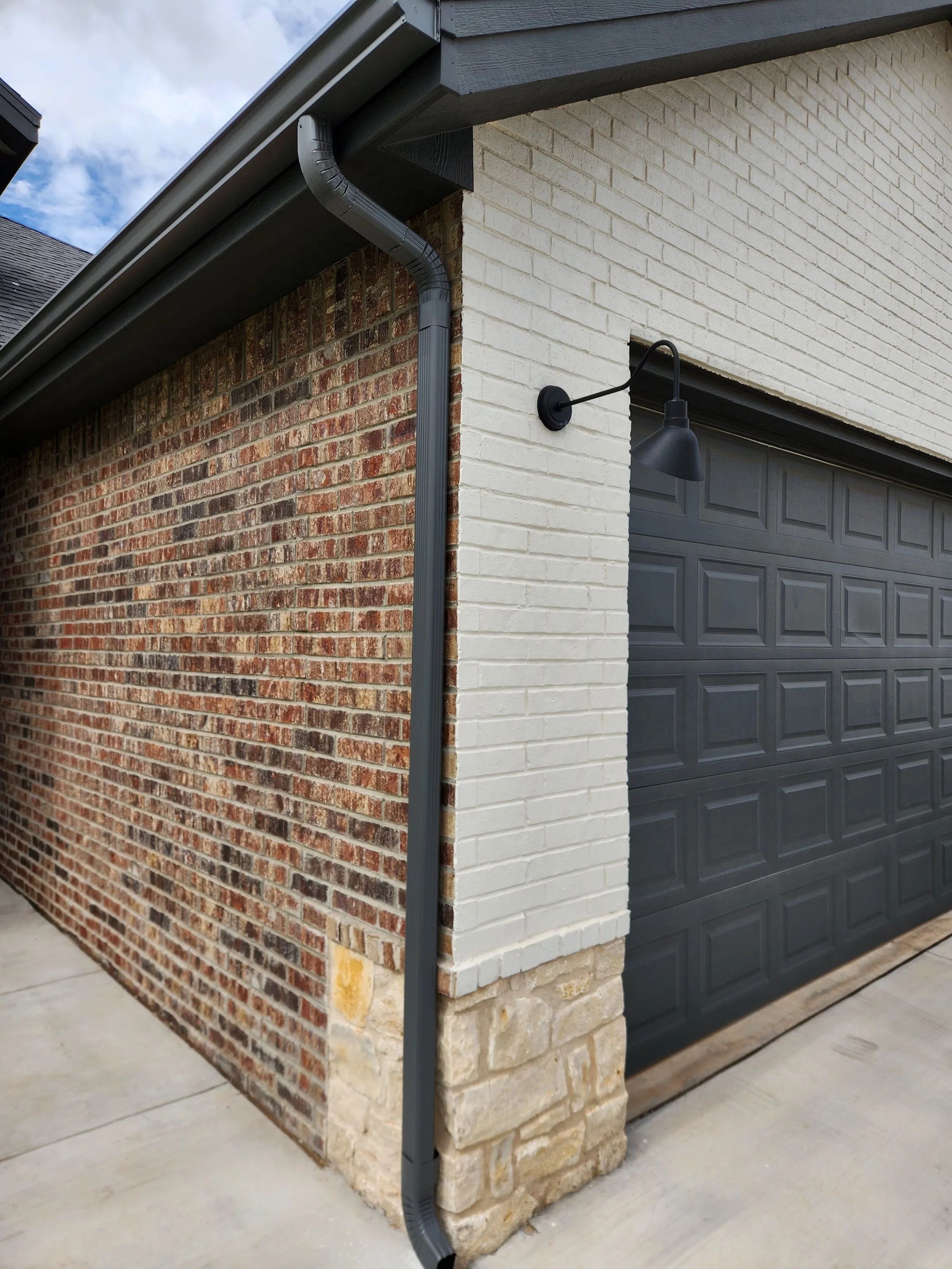 Corner of a house with brick and white painted brick walls, a black garage door, a black outdoor wall light, and a black gutter with downspout, on a concrete floor.