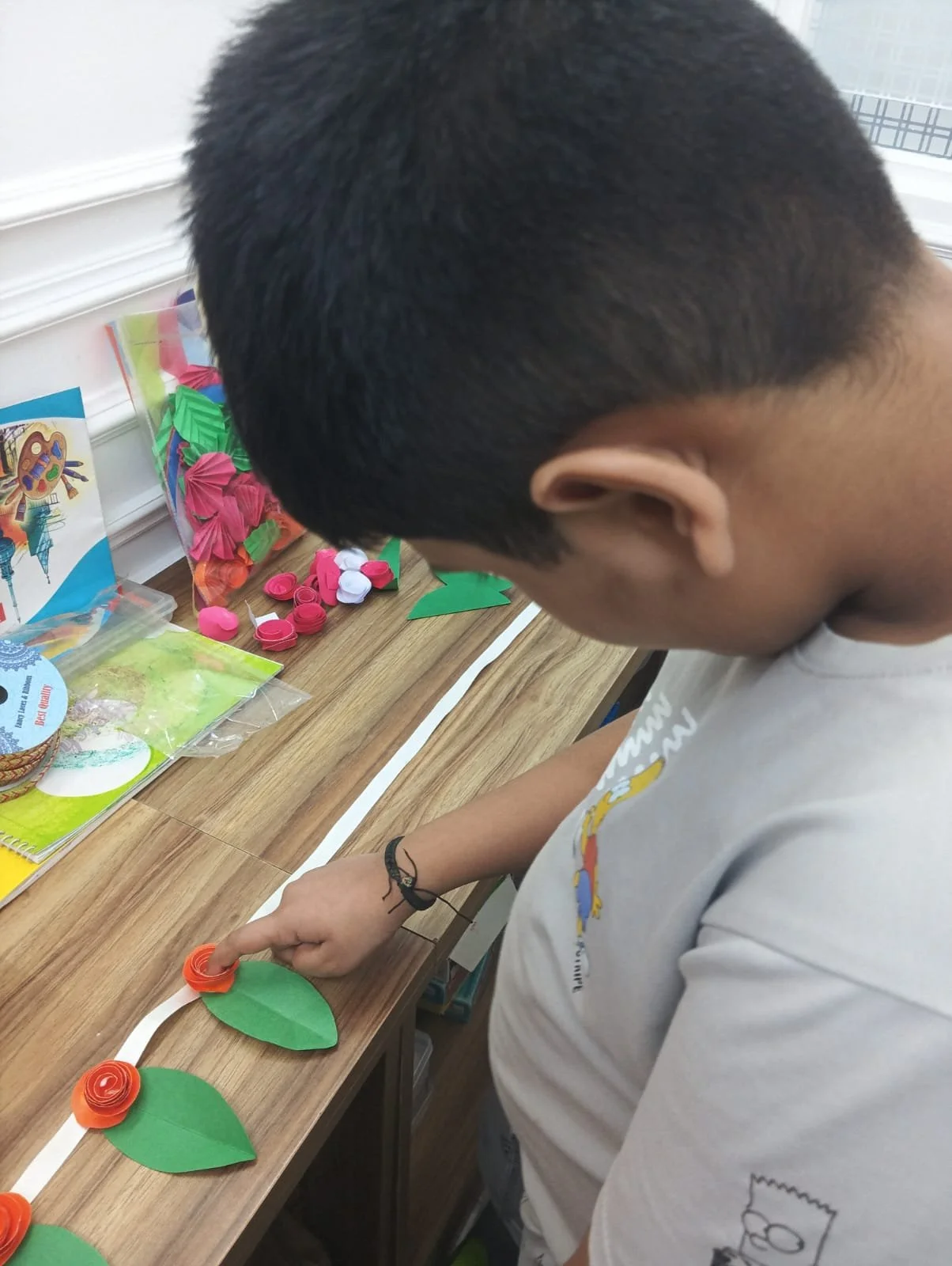 A boy with short black hair and a gray shirt with a cartoon character on sleeve, working on a craft project at a table. There are colorful paper crafts, including flowers made from paper and some completed paper flowers, on the table.