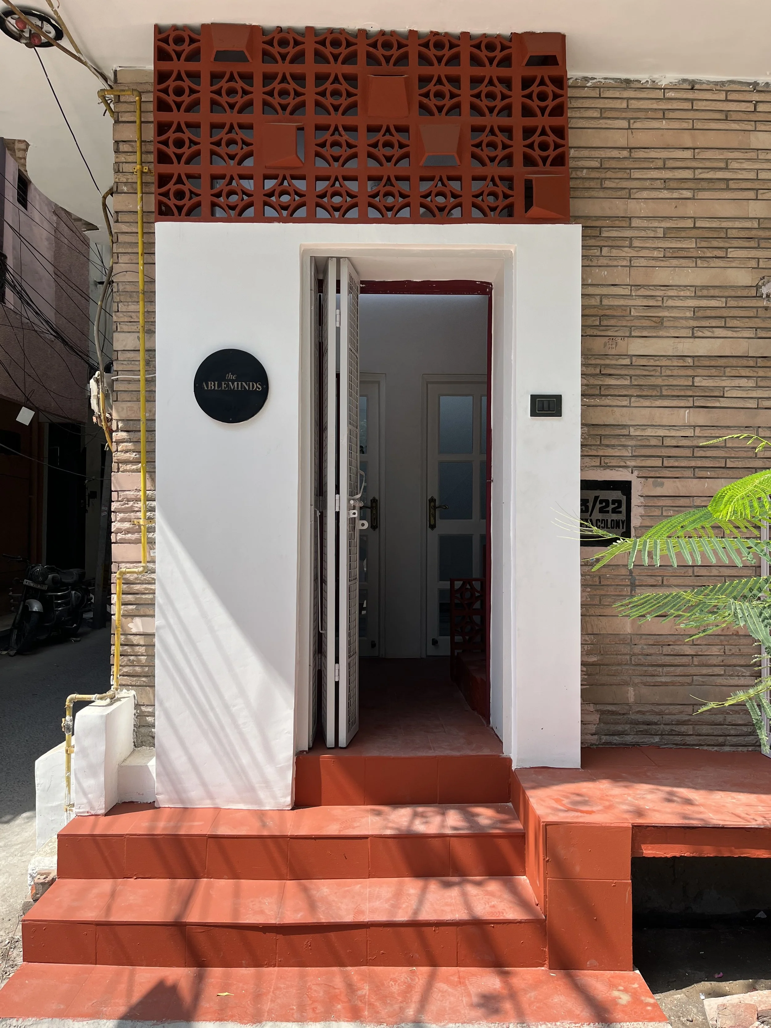 Entrance to a building with white walls, brown brick wall to the right, and red tiled steps leading up to a door. The door is open, revealing an interior with white walls and two windows. A round black sign with gold lettering reads 'the ABLEMINDS' is mounted on the wall to the left of the door. The front features a decorative red concrete screen on top of the wall.