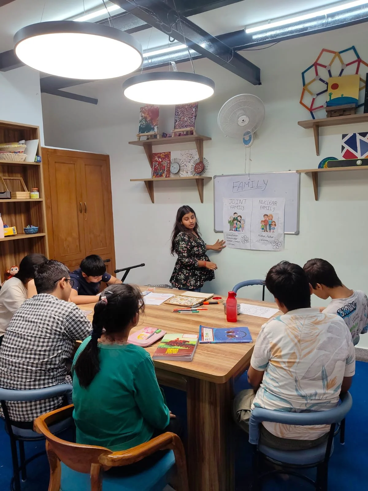 A classroom with students seated around a wooden table, listening to a female teacher standing in front of a whiteboard illustrating family relationships. The classroom is decorated with artwork, books, and shelves, and has bright lighting and ceiling fans.