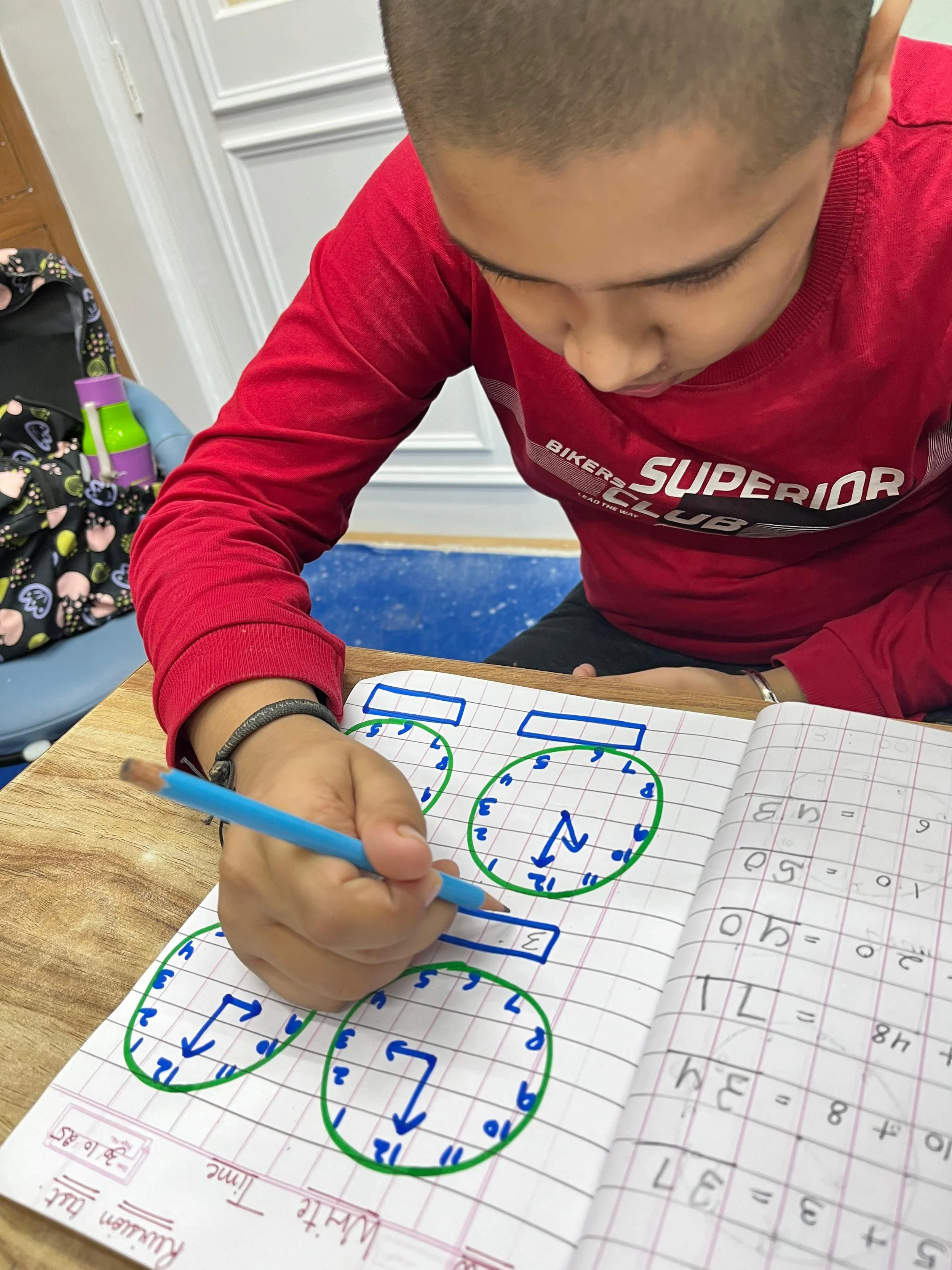 A person in a red long-sleeved shirt is writing mathematical diagrams in a notebook with a blue pen. The notebook contains multiple green circles with blue number sequences and arrows, along with some workbook pages featuring mathematical problems and equations. The scene is set on a wooden table with a backpack nearby.