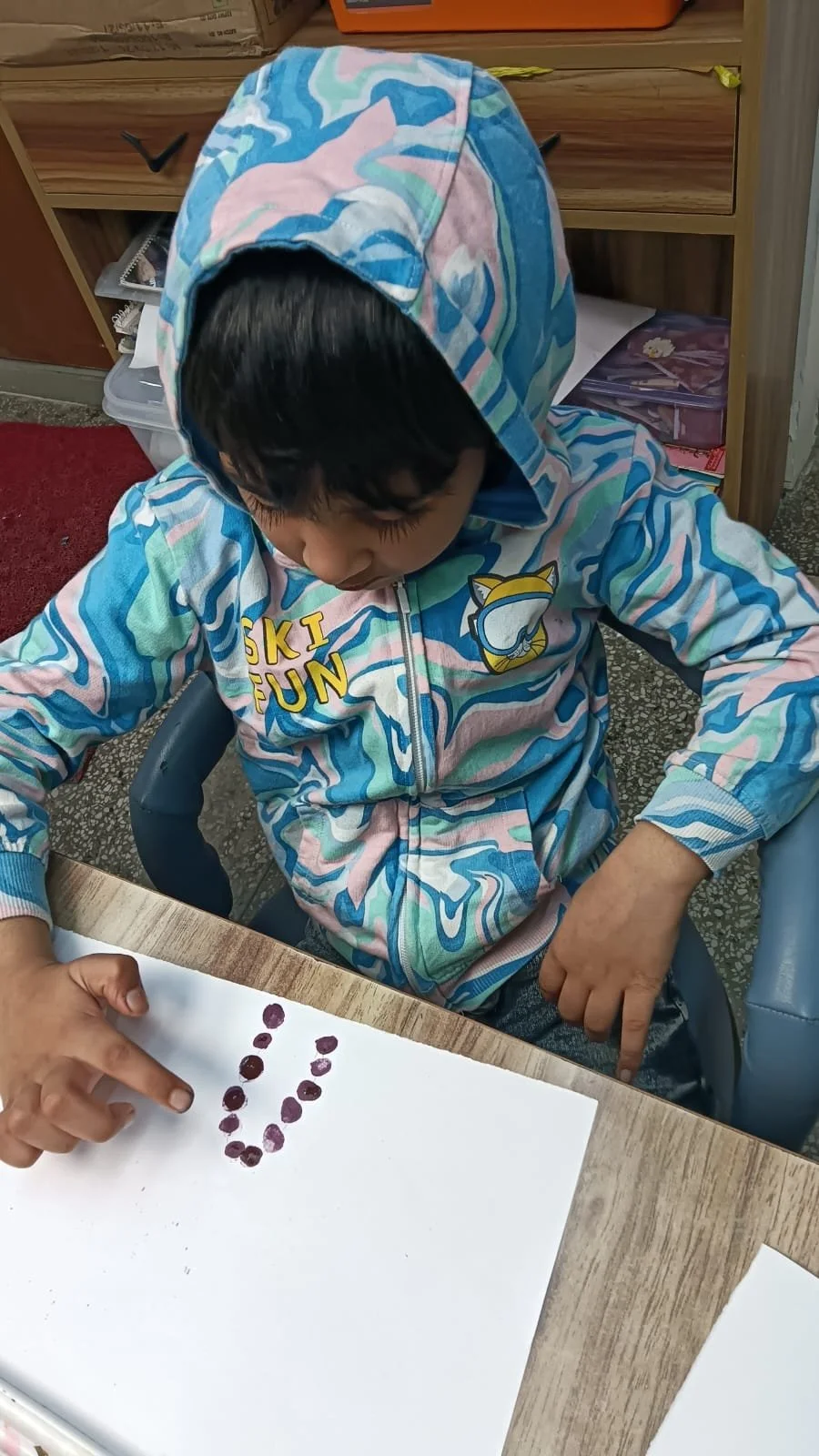 A young child wearing a colorful hoodie with a 'Ski Fun' logo and a cartoon cat on it, pointing at a white sheet of paper with purple paint dots arranged in a smiley face.