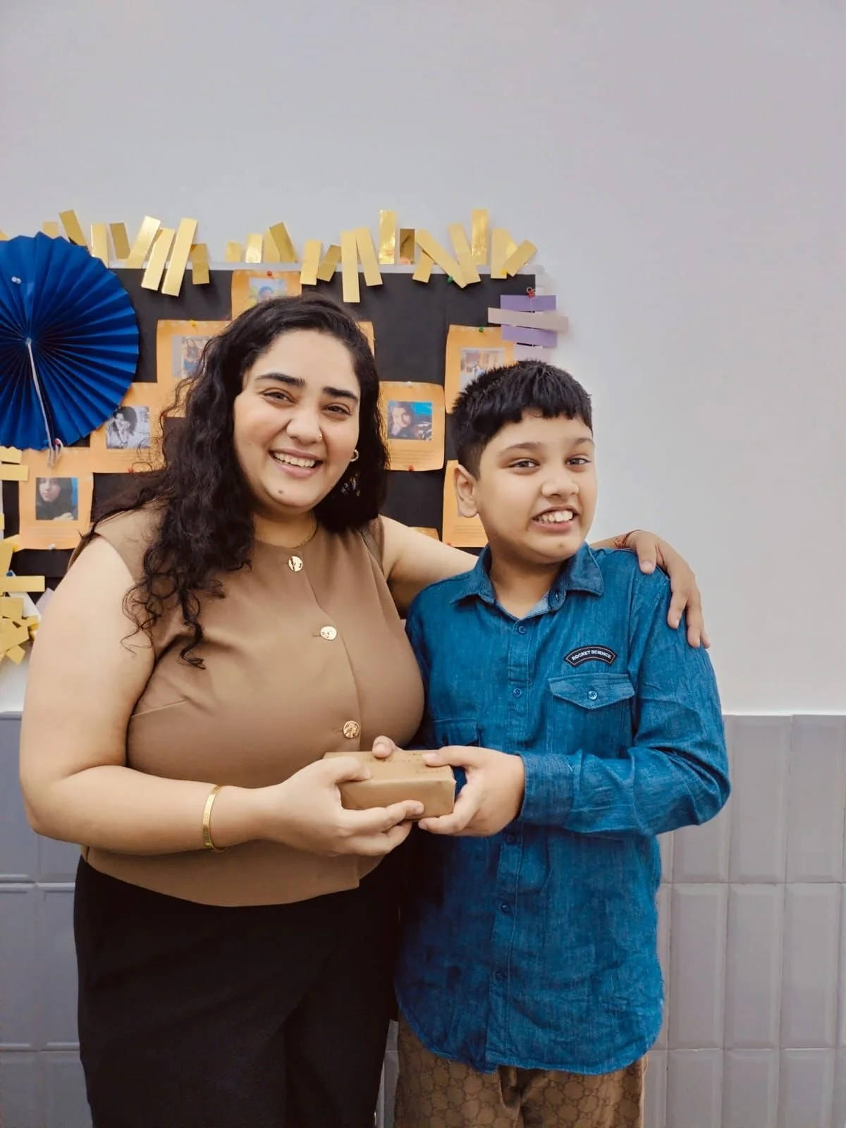 A woman and a boy are standing together, smiling at the camera. The woman has long dark hair and is wearing a beige sleeveless top. The boy has short black hair and is wearing a blue button-up shirt. They are indoors, with a decorated black bulletin board with photos and yellow paper strips in the background. The woman is holding a small wrapped gift.
