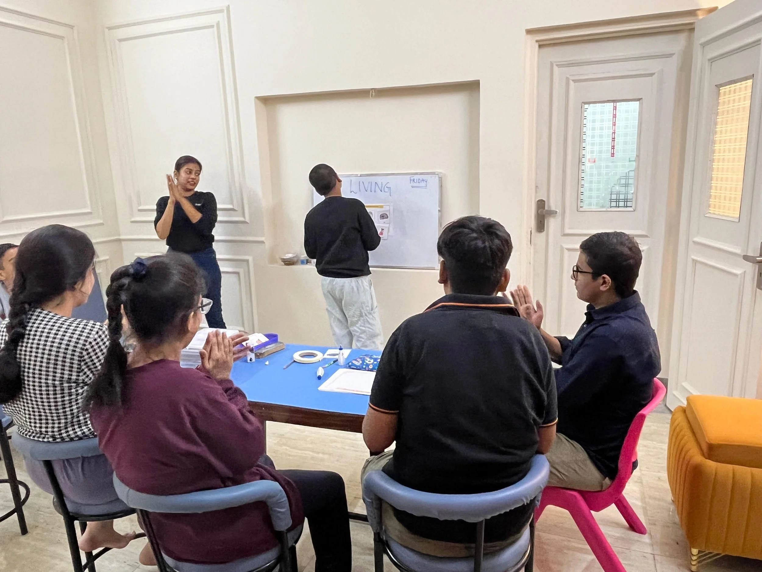 A group of people seated around a table in a room with white paneled walls, facing a woman standing near a whiteboard. The woman appears to be speaking or teaching, and a boy is writing on the whiteboard that has the word 'LIVING' written on it, along with some other notes. The room has a door and a window with blinds.