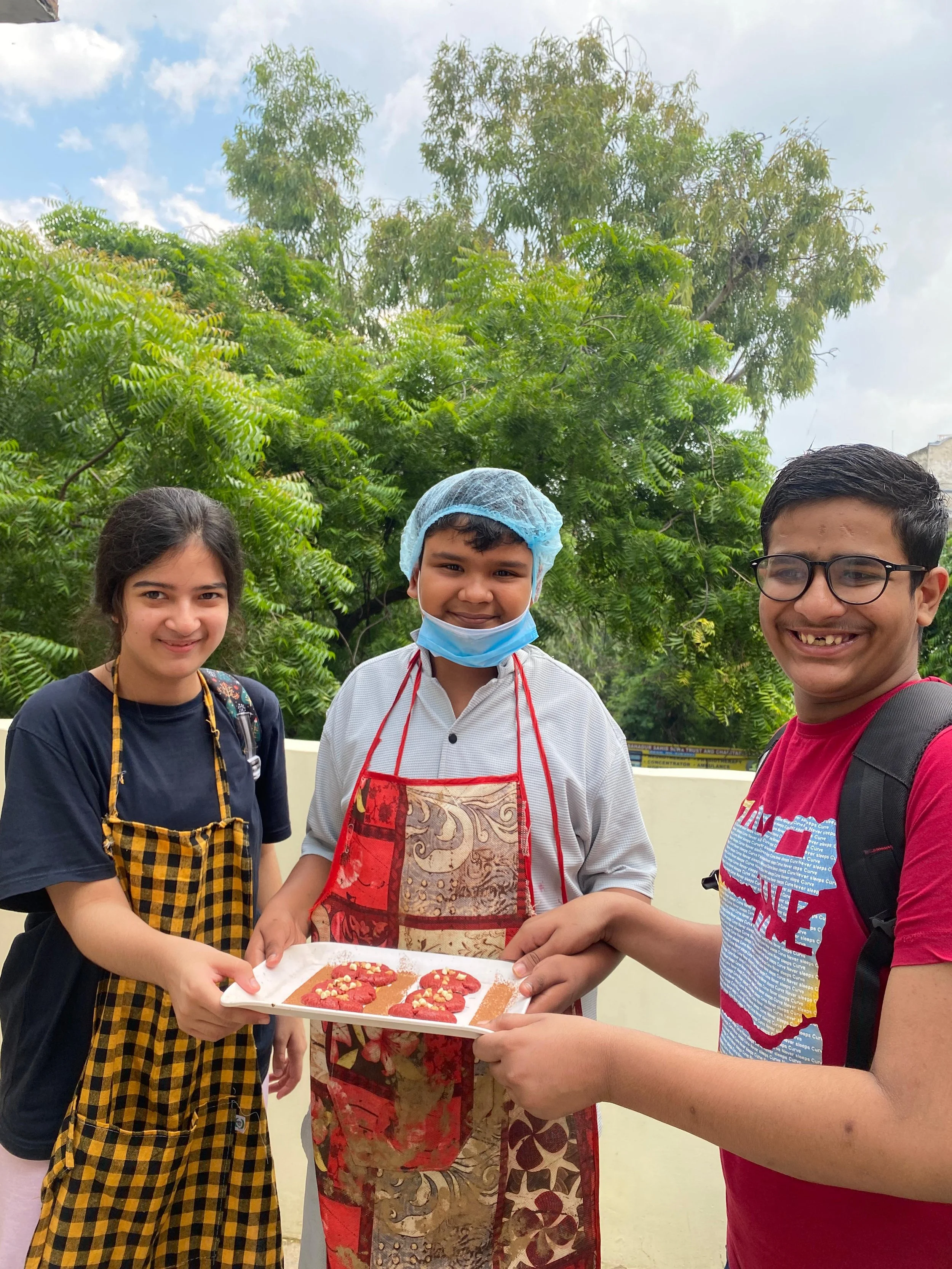 Three people smiling outdoors, one of them wearing a hairnet and an apron, holding a tray of decorated cookies; another person is wearing glasses and a red T-shirt, and a young woman with long dark hair and a black top. There are green trees and a cloudy sky in the background.