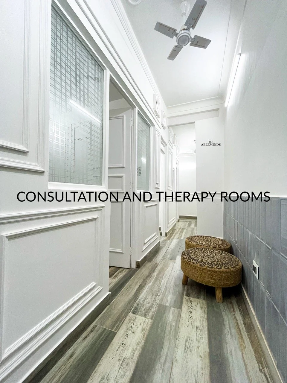 A hallway with white walls and decorative molding, two small upholstered stools with patterned cushions, a ceiling fan, and a sign reading 'the ABLEMINDS'. Text overlay states 'Consultation and Therapy Rooms'.