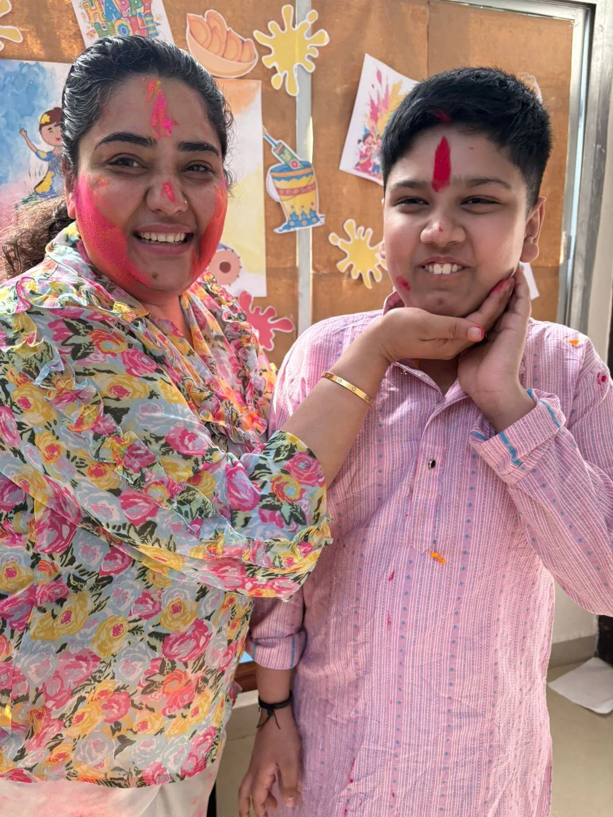 A woman and a boy celebrating Holi, with colored powder on their faces and clothes, standing in front of a festive wall with colorful posters and drawings.
