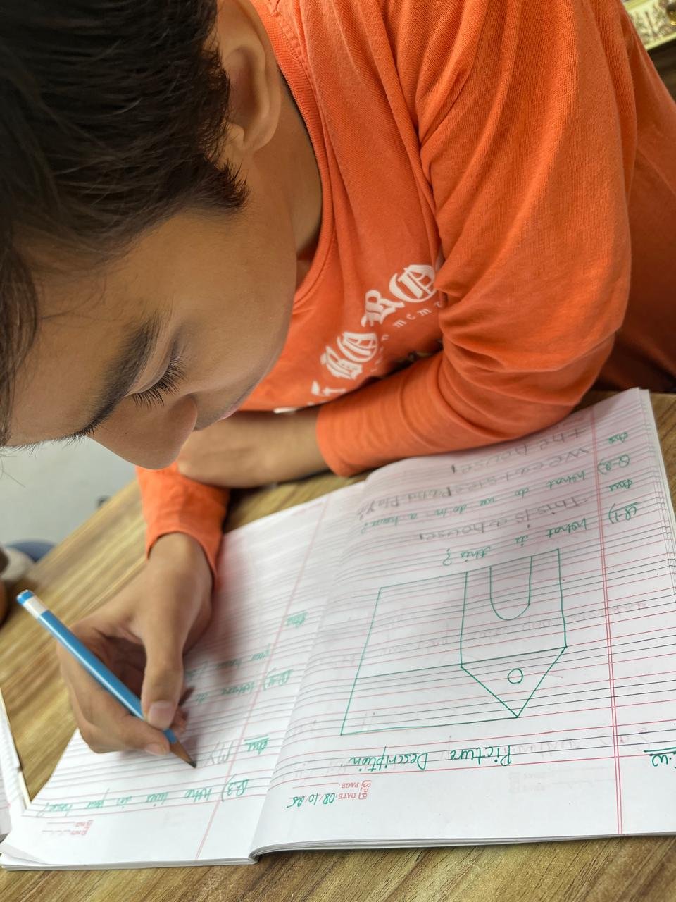 A young boy in an orange shirt writes in a notebook with a blue pen, sitting at a wooden desk.