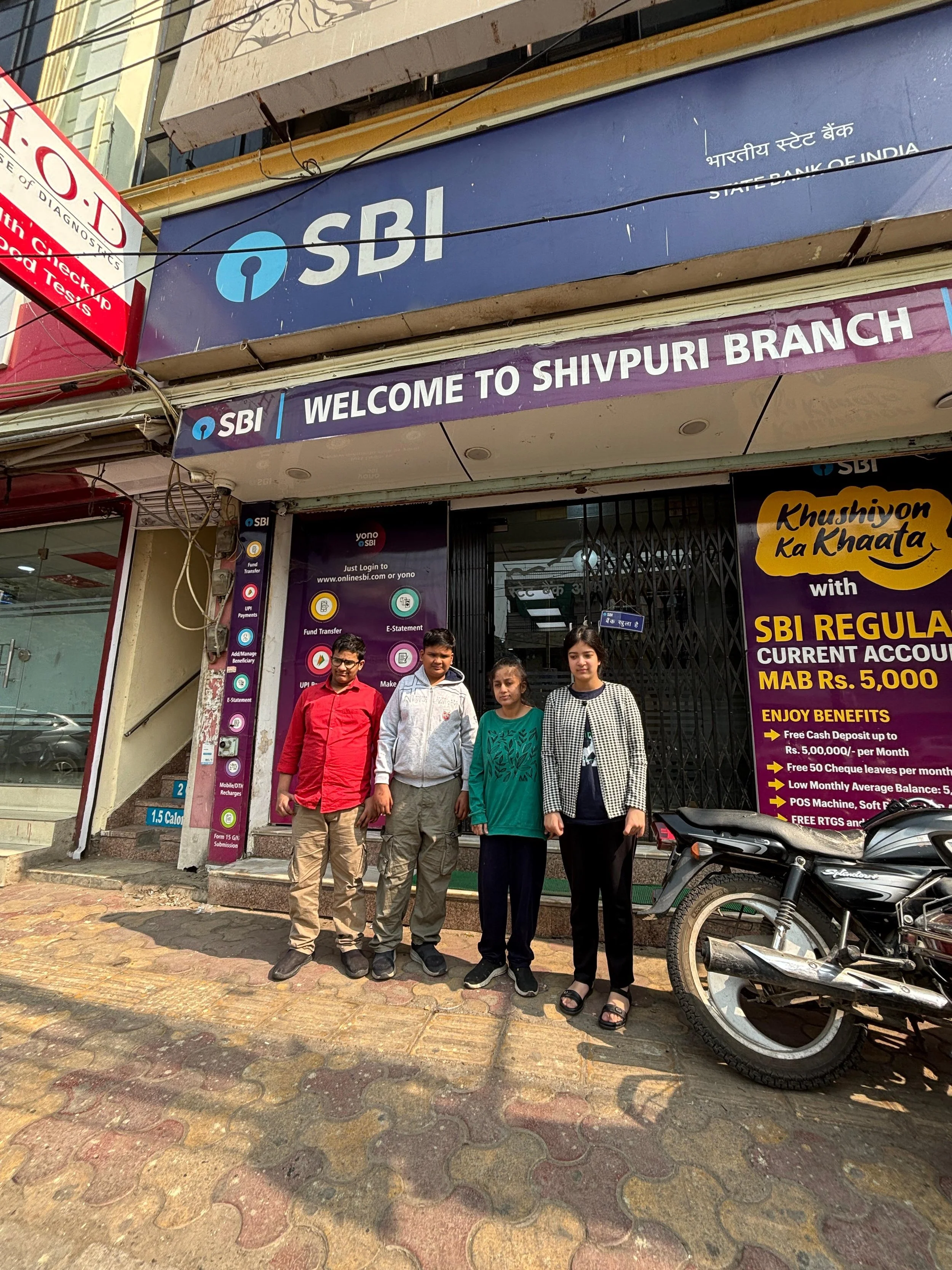 Four young people standing in front of an SBI bank branch on a busy street, with a motorcycle parked nearby. The bank sign above reads "WELCOME TO SHIVPURI BRANCH" and features promotional banners.