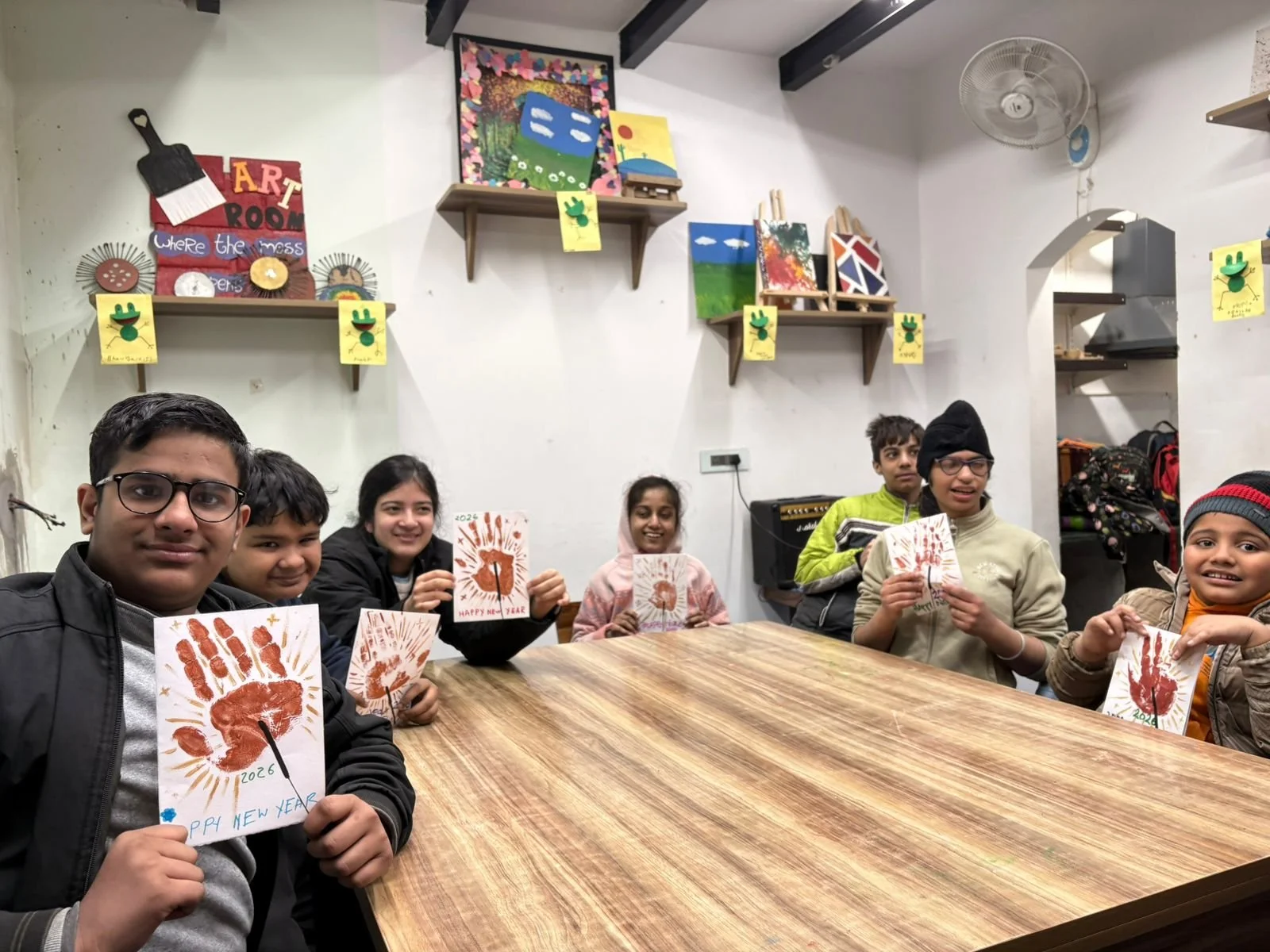 A group of children sitting around a wooden table in a room, holding up New Year craft cards with handprint and paint designs. The room has art on the walls and shelves with artwork, and children appear happy and proud of their projects.