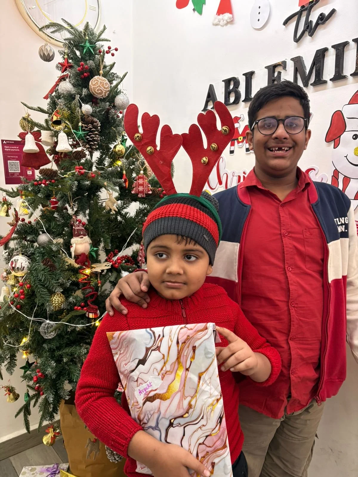 Two boys standing in front of a decorated Christmas tree. The boy in the front is wearing a red sweater and a gray and red beanie with reindeer antlers, holding a gift. The taller boy behind him is wearing glasses, a red shirt, and a jacket, smiling. The background has Christmas decorations and a sign that says "ABLEMIR".