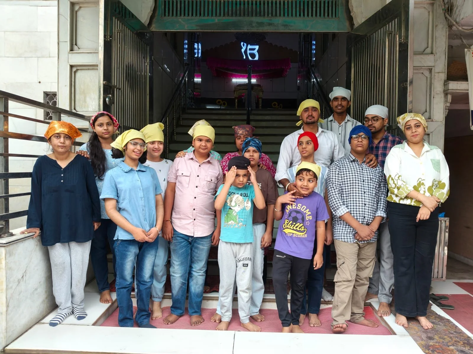 Group of children and adults standing on a staircase inside a building, some wearing colorful head coverings, in front of a temple or religious site with a decorated canopy and symbols in the background.