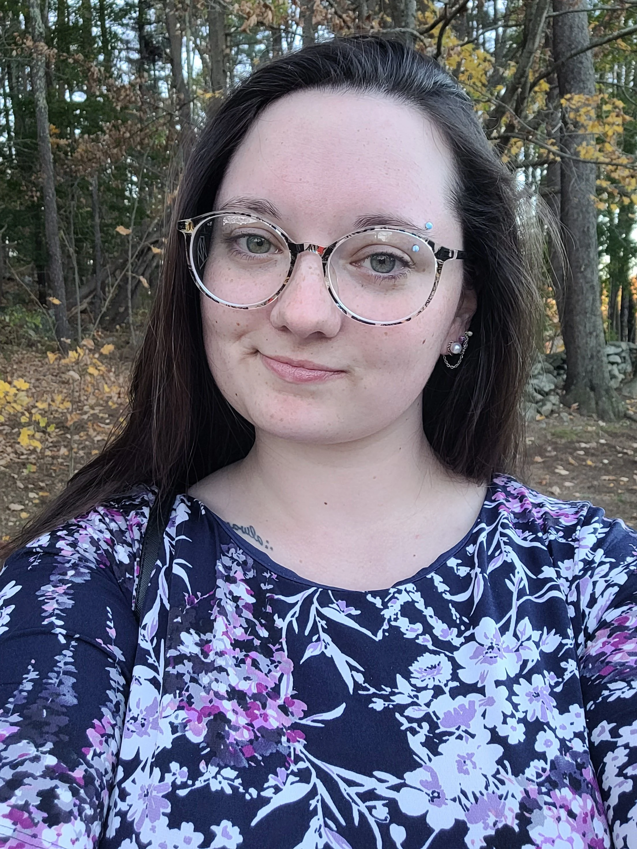 A young woman with long dark hair, wearing glasses with a tortoiseshell frame, standing outdoors in a wooded area with trees and fallen leaves, taking a selfie.