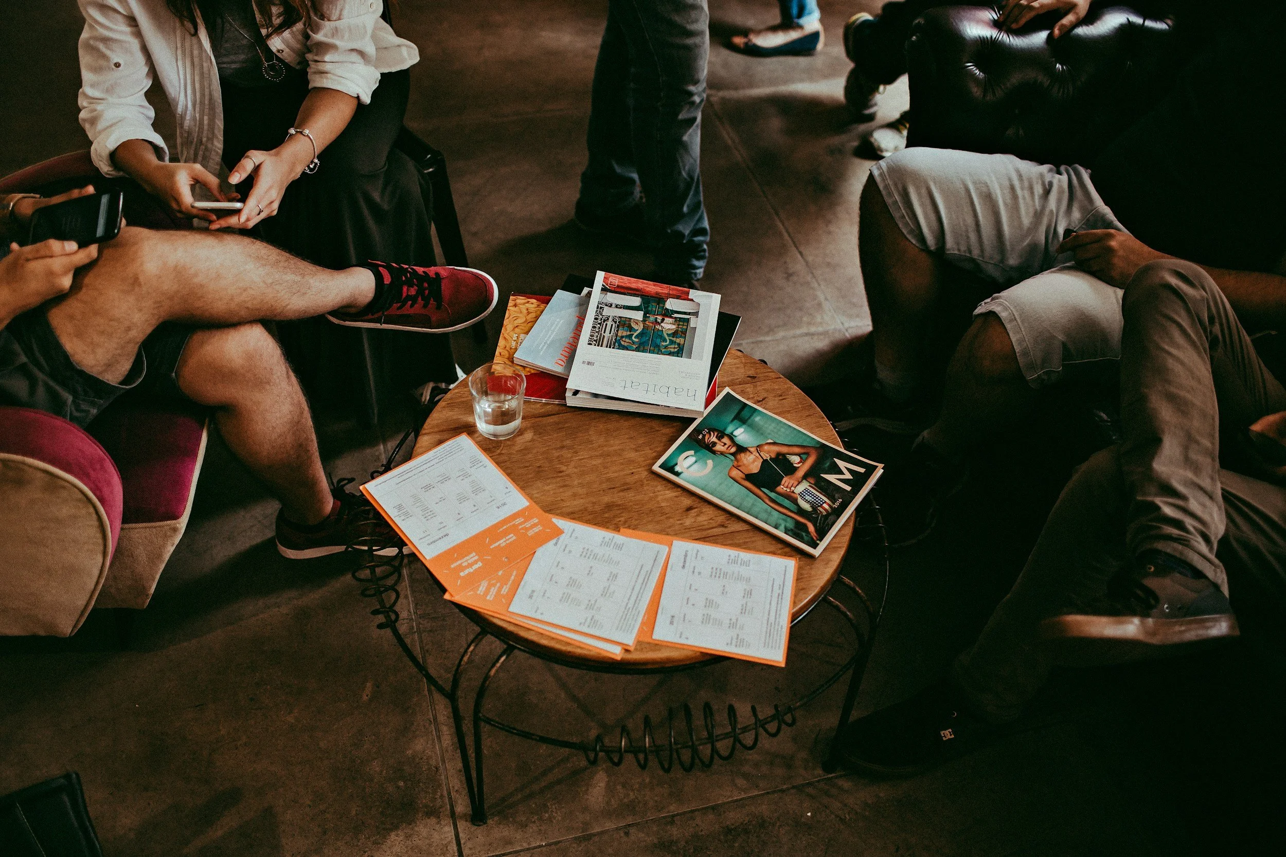 A group of people sitting and standing around a coffee table with magazines, menus, and a glass of water on it, in a casual indoor setting.