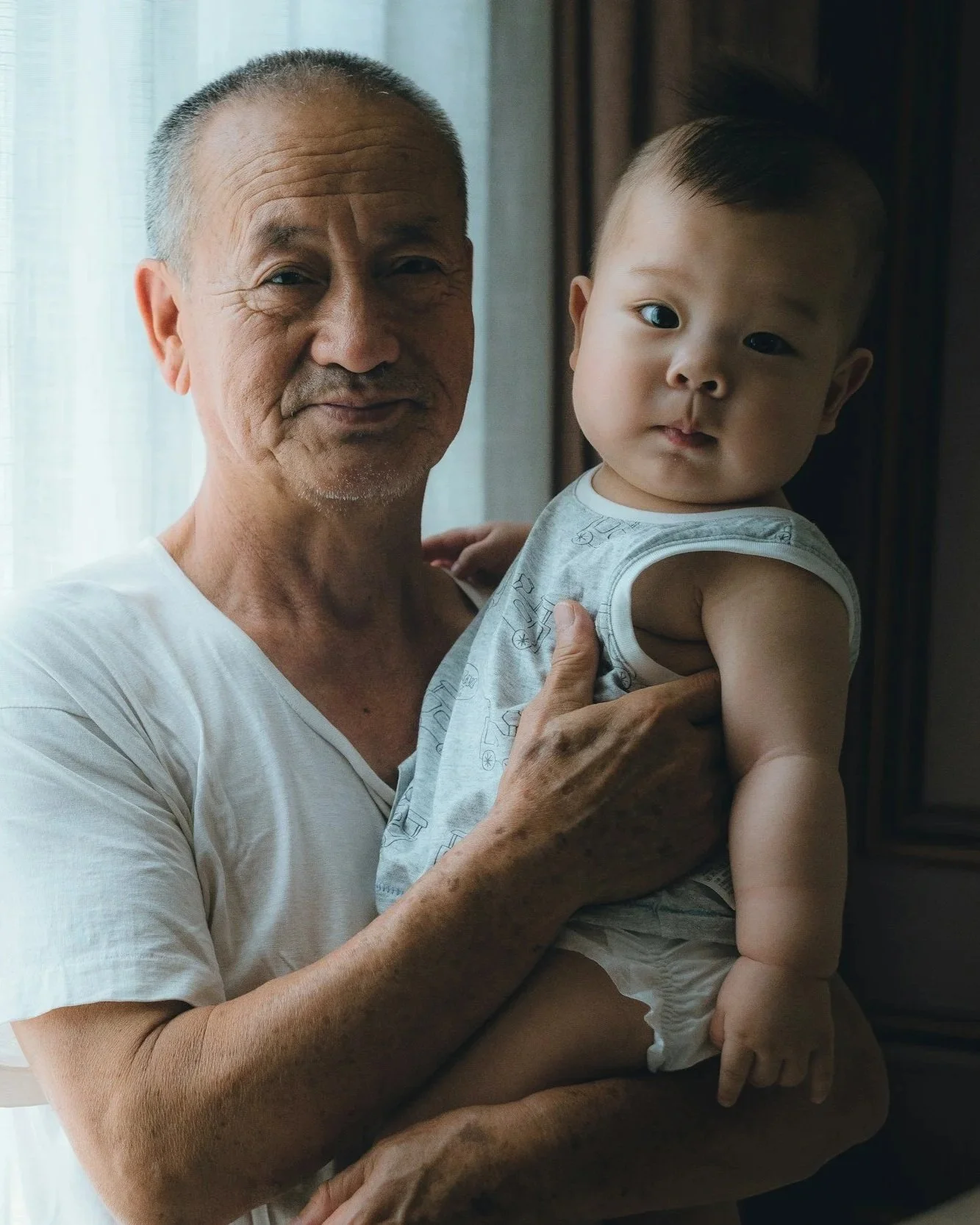 An elderly man with short gray hair holding a young child with short dark hair in his arms, standing near a window with light-colored curtains.