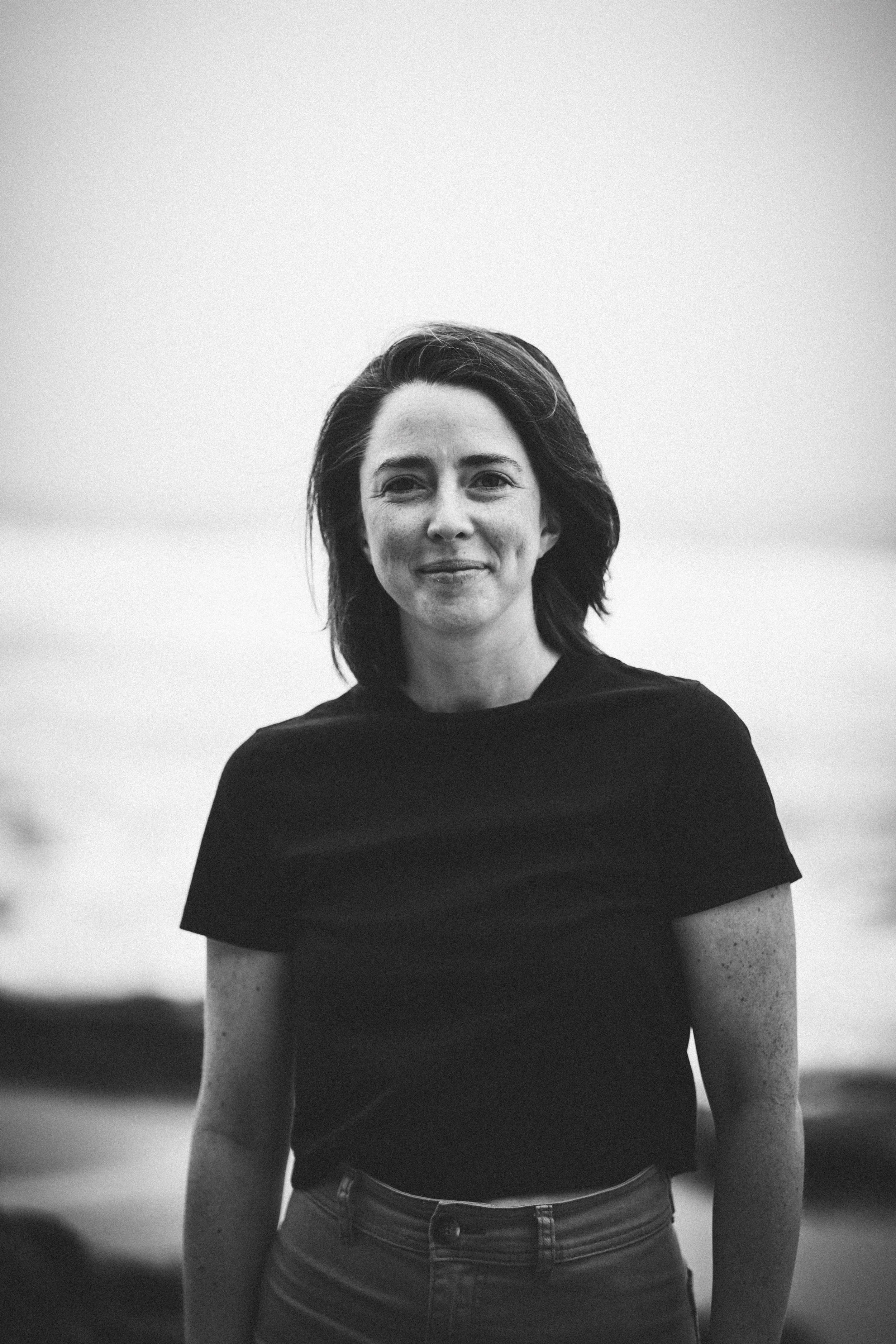 Black and white photo of a woman standing outdoors, smiling gently, wearing a dark t-shirt and jeans. The background is blurred, suggesting an open outdoor setting.