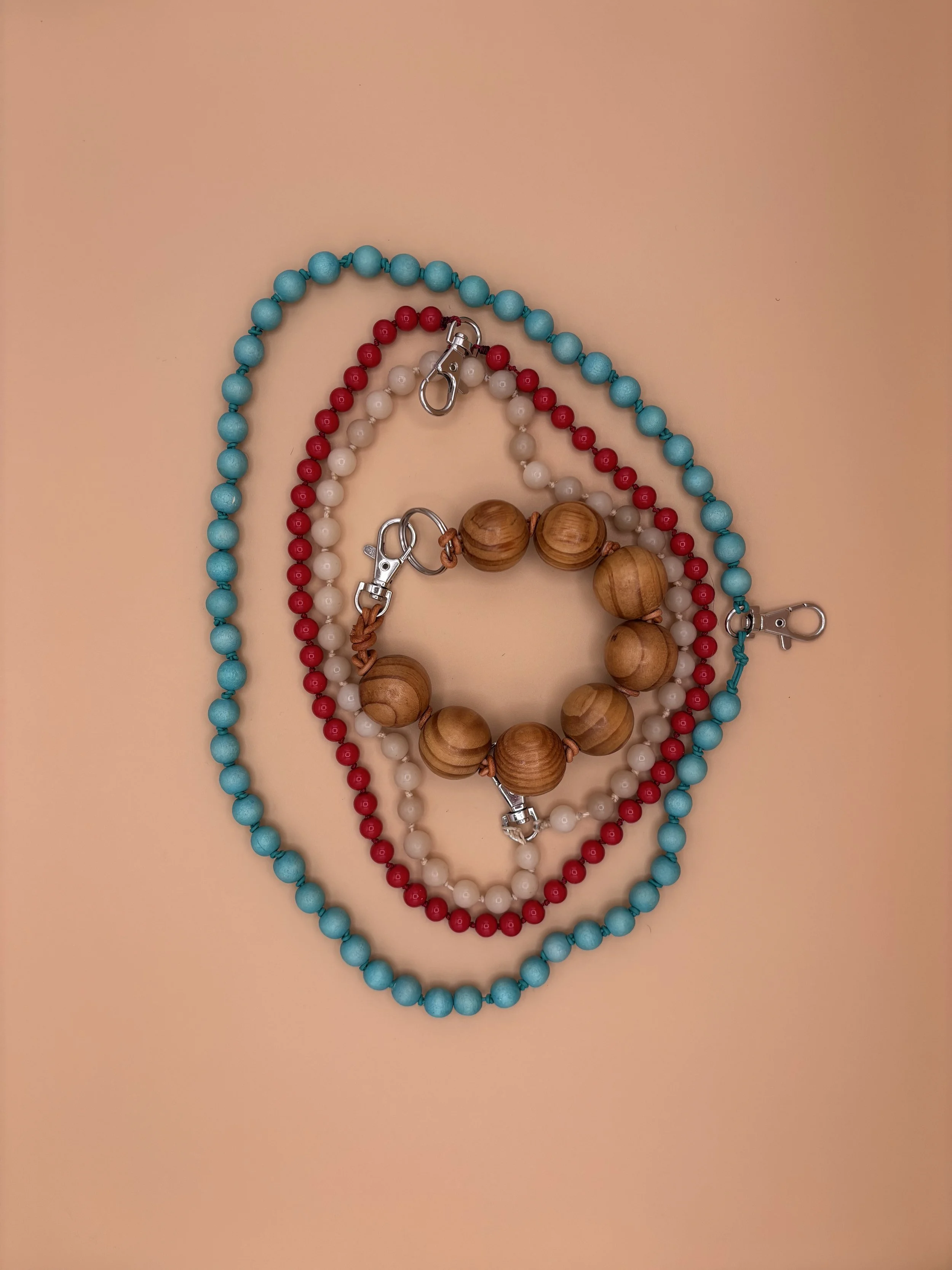 Three beaded necklaces in blue, red, and white with large wooden beads, laid out on a peach-colored background, with clip hooks attached to the necklaces.