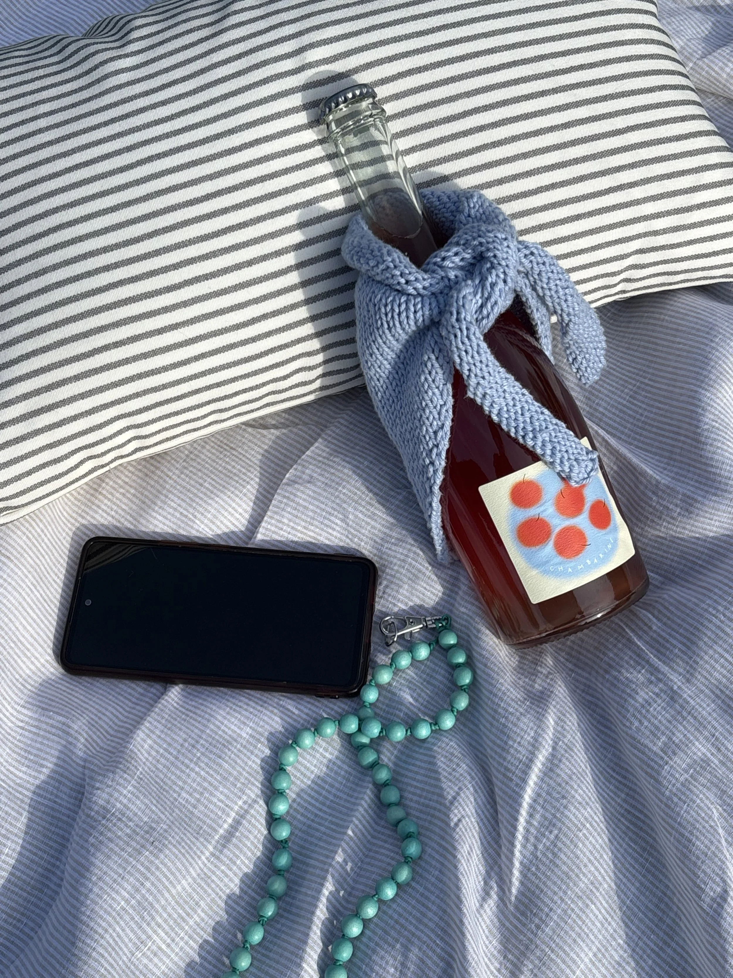 A collection of personal items laid out on a striped fabric surface, including a smartphone, a set of keys, a teal beaded necklace, a bottle of sparkling beverage with a label, a dark blue cloth or garment, and a small round object.