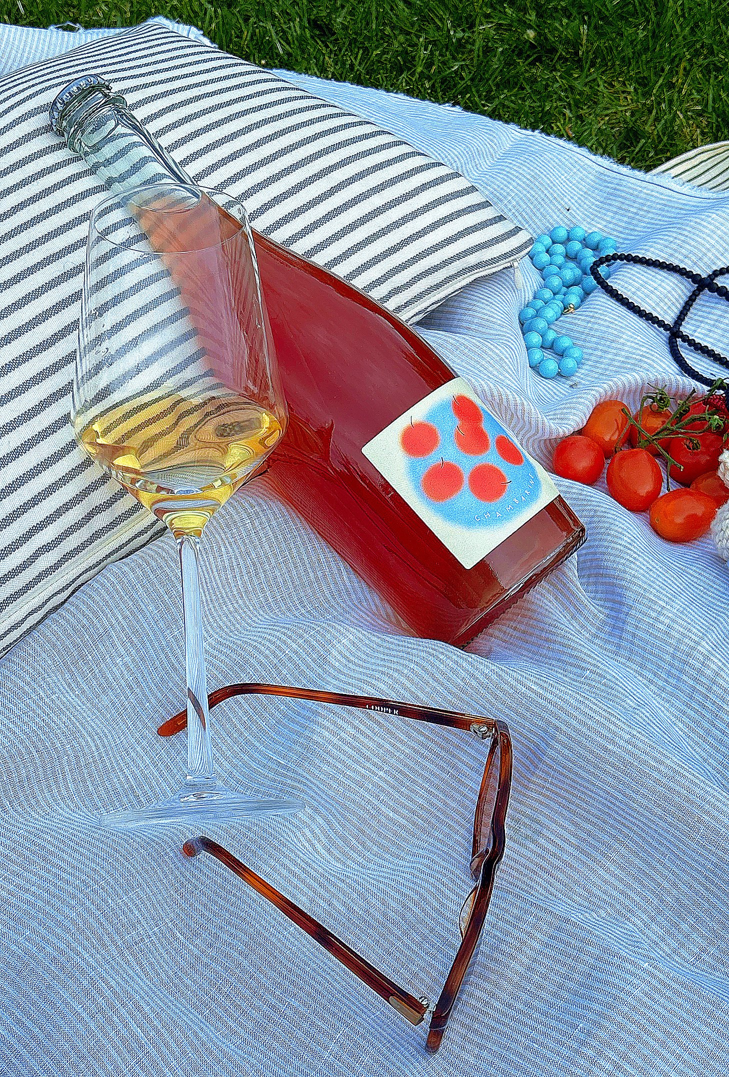 A wine glass filled with white wine, a bottle of rosé wine with a label featuring red tomatoes, and an assortment of tomato and blue beads on a white and blue striped cloth outdoors.