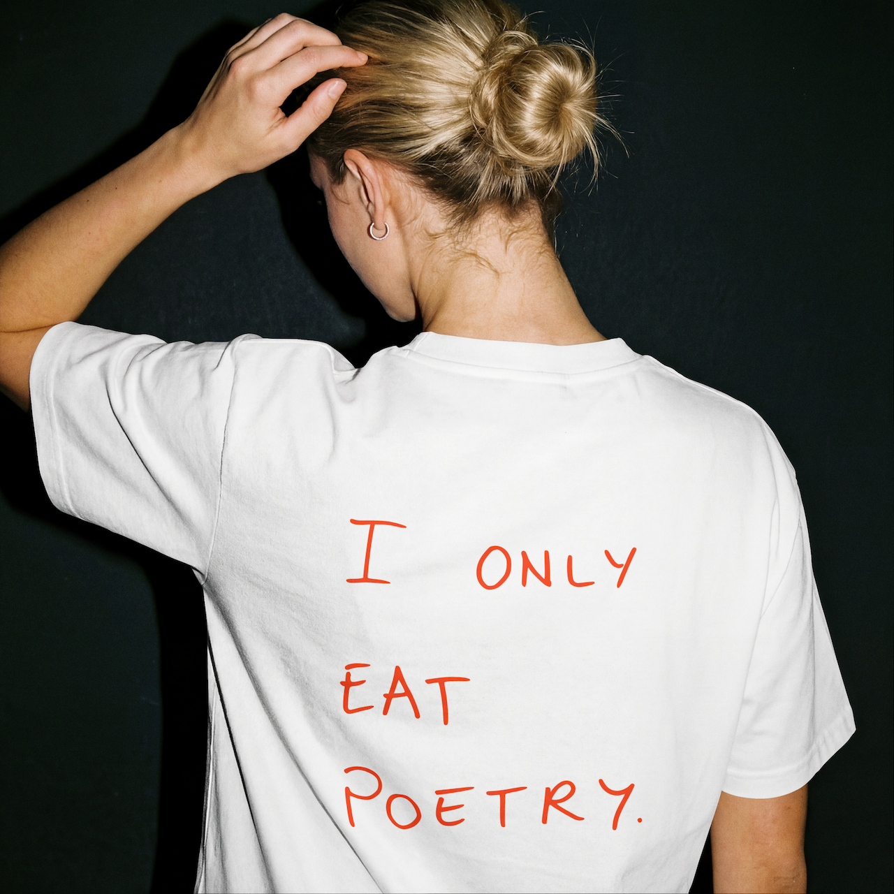 Back of a woman wearing a white T-shirt with red handwritten text that reads 'I ONLY EAT POETRY,' with her hand resting on her head and a black background.