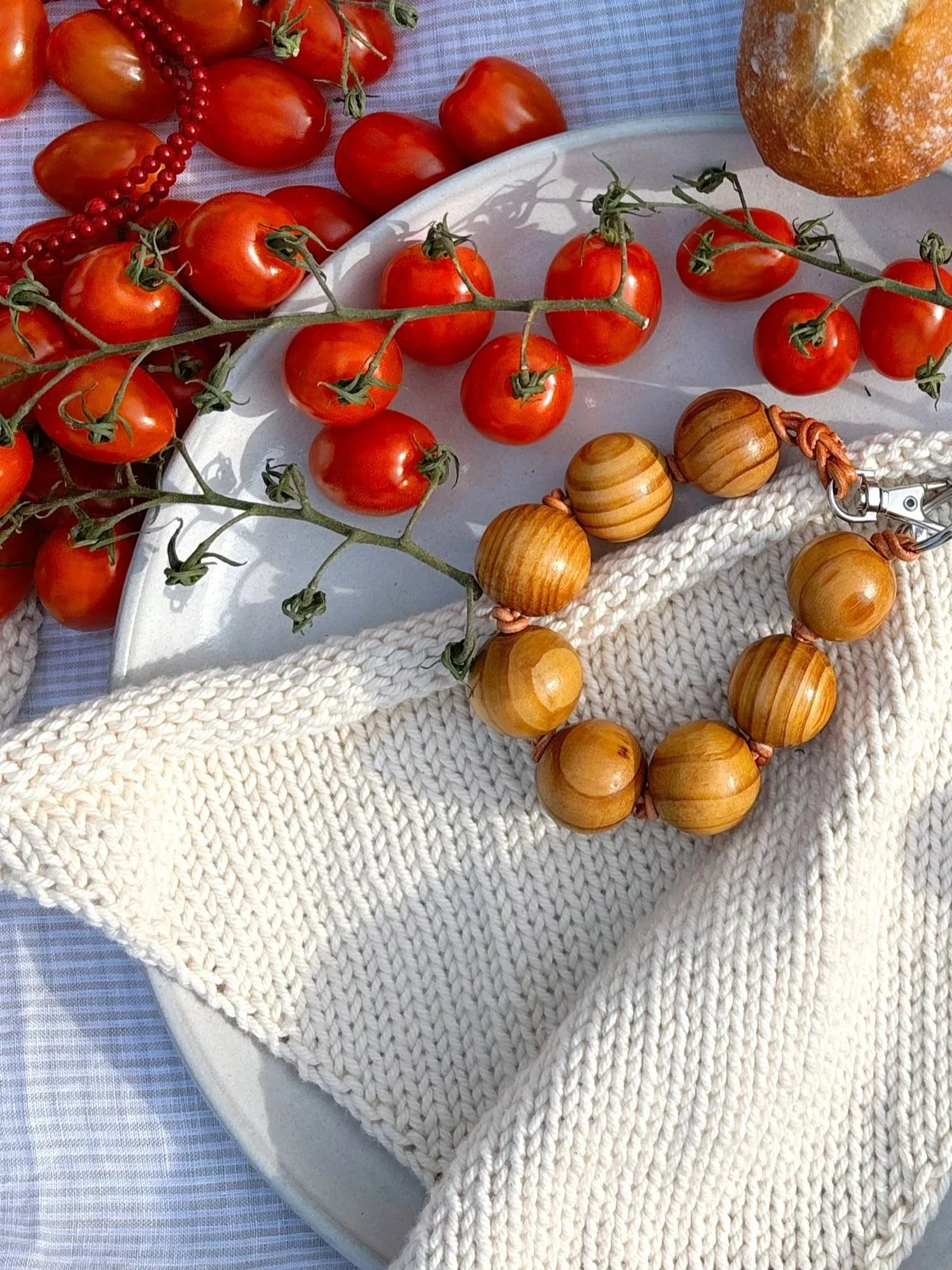 A white platter with fresh cherry tomatoes on the vine, a loaf of bread, a string of wooden beads, a knitted white cloth, and a red beaded necklace, all arranged on a light-colored tablecloth.
