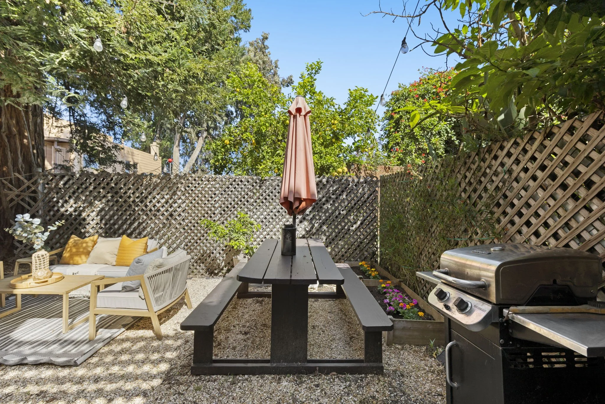 An outdoor patio with a picnic table, a grill, a cushioned seating area, and a flower bed, surrounded by trees and a lattice fence, under a blue sky with string lights hanging overhead.