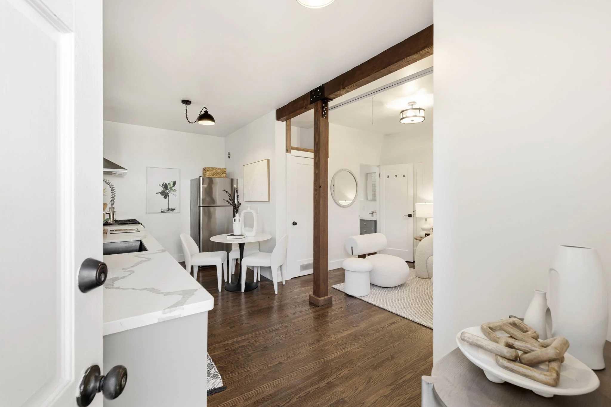 Living room and kitchen area in a modern home with white walls, dark wood flooring, and minimalist furniture. The living room includes white chairs, a round ottoman, and a small side table with decorative vases. The kitchen features a stainless steel
