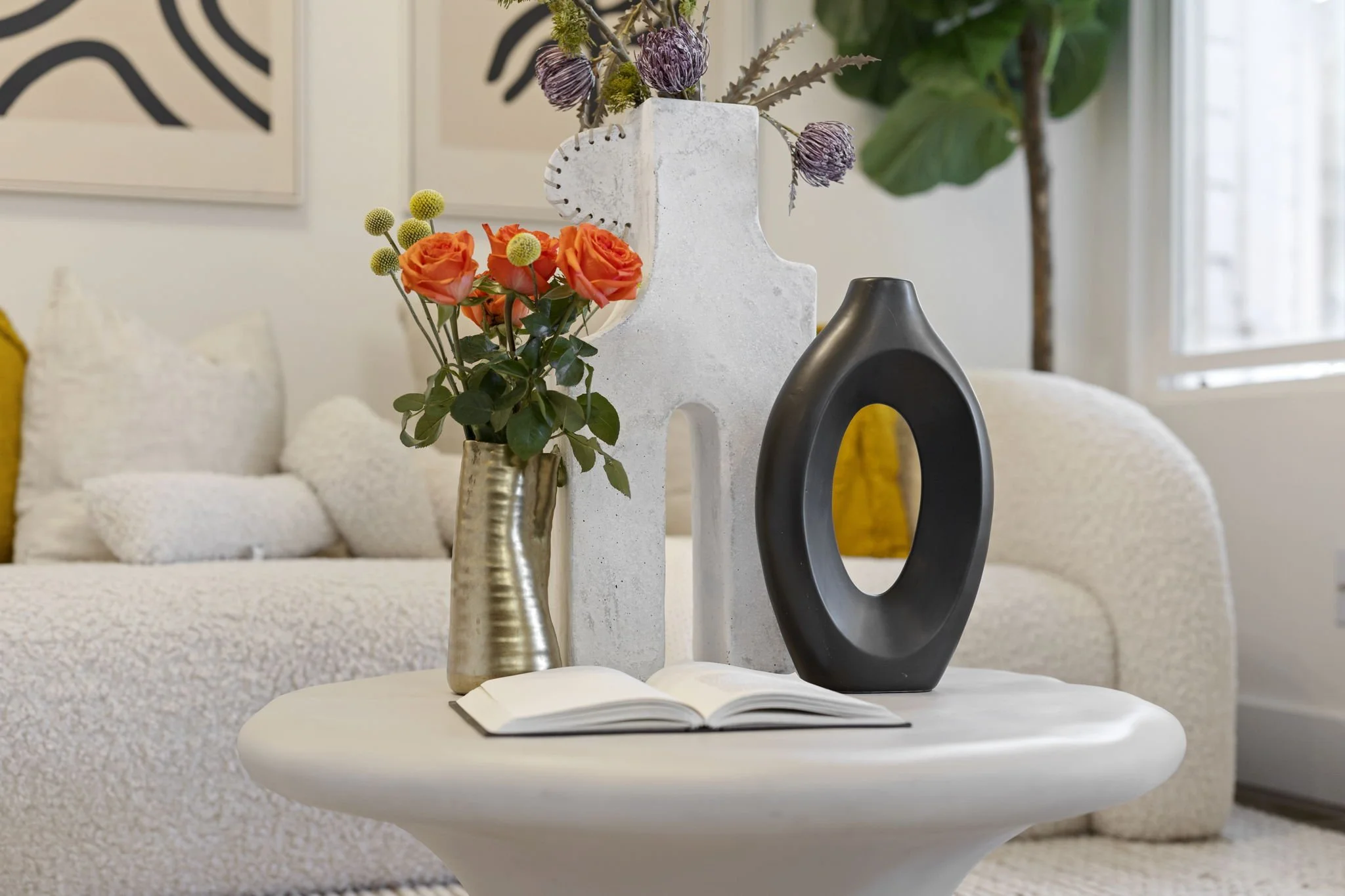 Decorative living room table with vases of flowers, a book, and abstract sculptures, with a white sofa and framed artwork in the background.