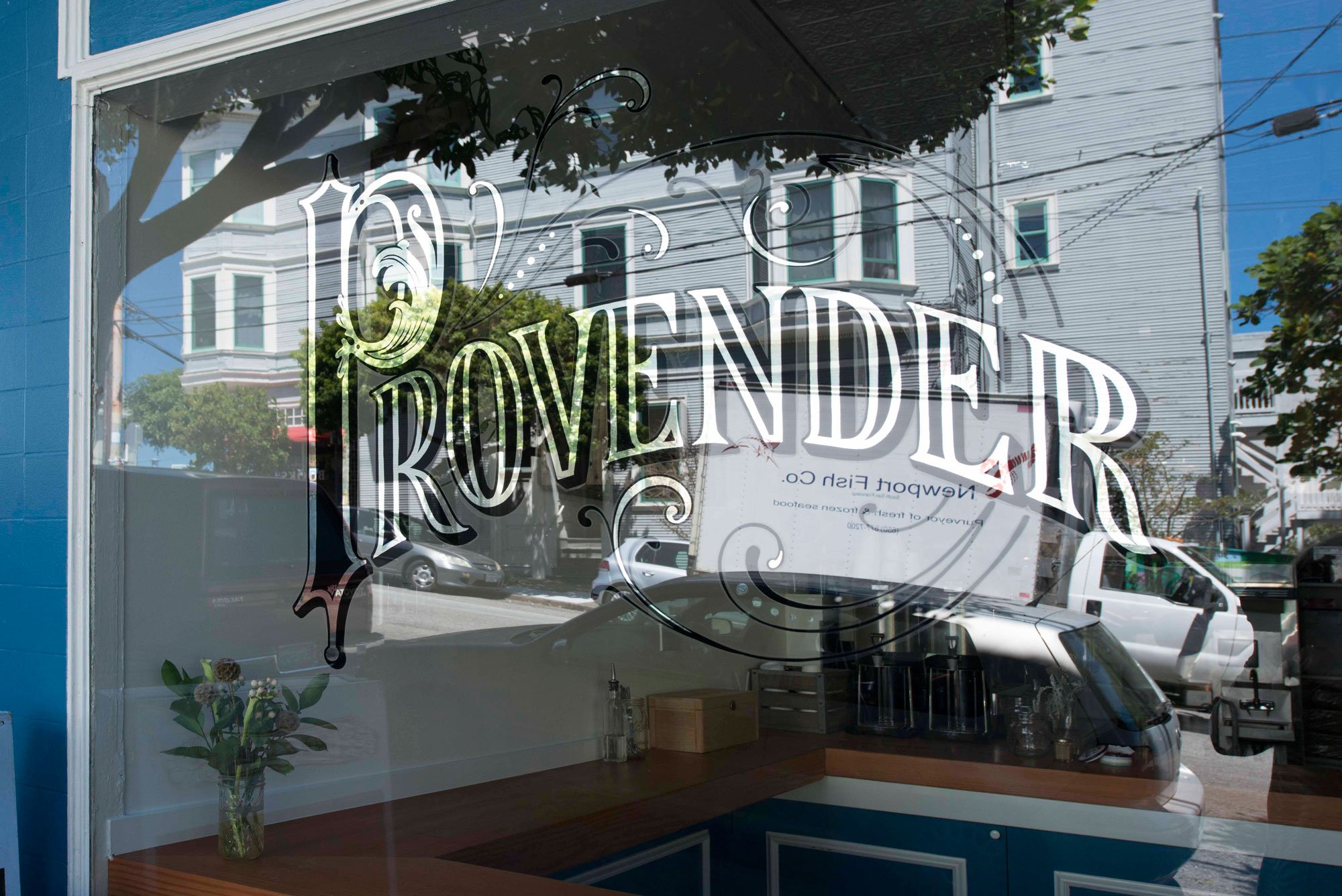 View through a glass window of a shop sign that reads 'The ROVENDER' with decorative swirls and a floral design, reflecting street and buildings outside.
