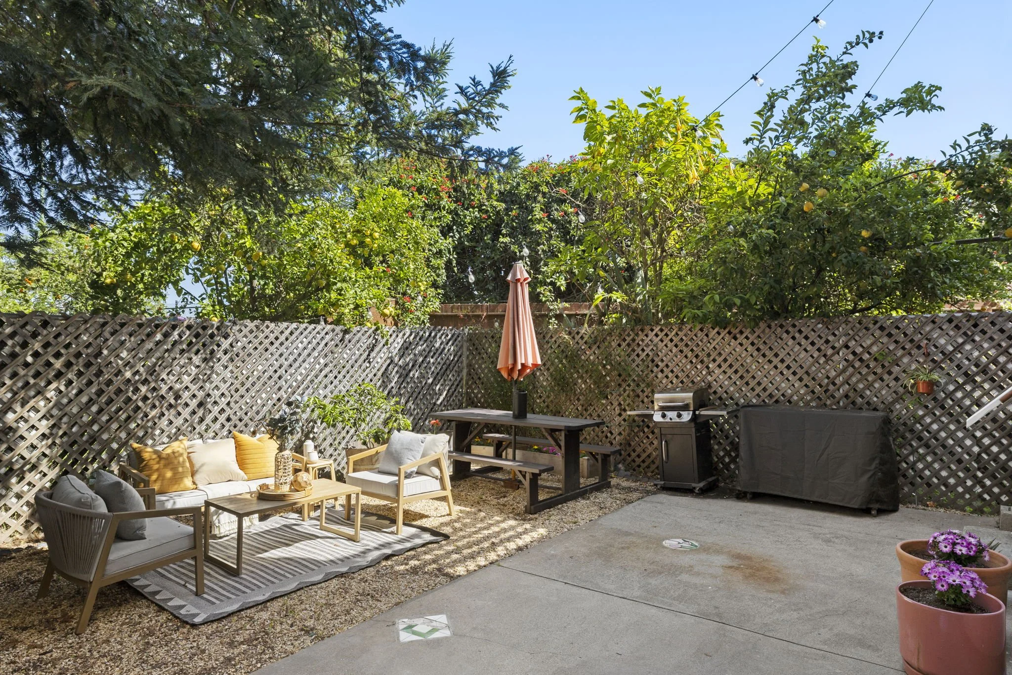 Backyard patio with outdoor furniture, a barbecue grill, potted flowers, and surrounded by a wooden lattice fence with trees and green foliage beyond.