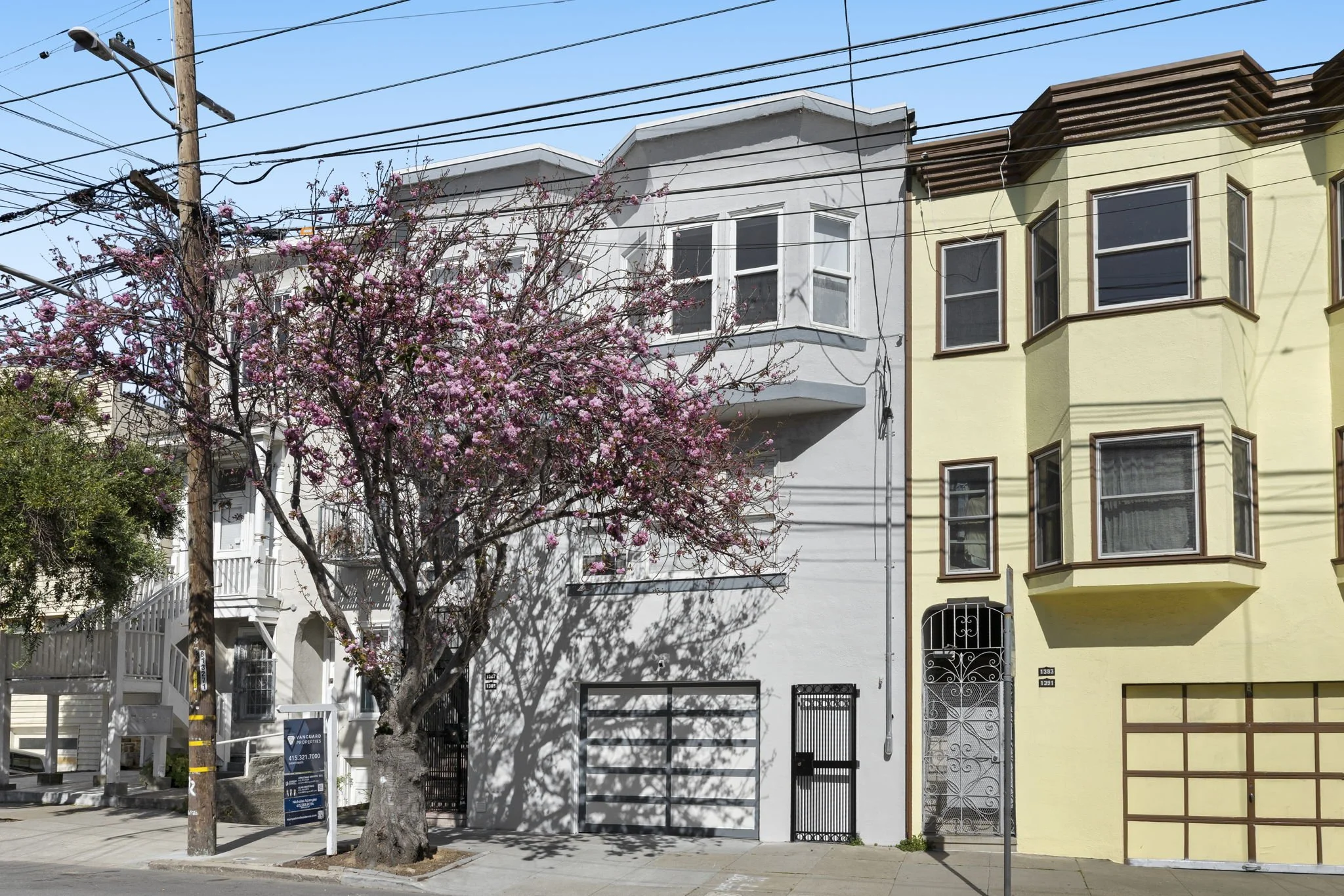 A white multi-story residential building with a small balcony, surrounded by a pink flowering tree, power lines, and a utility pole in front.