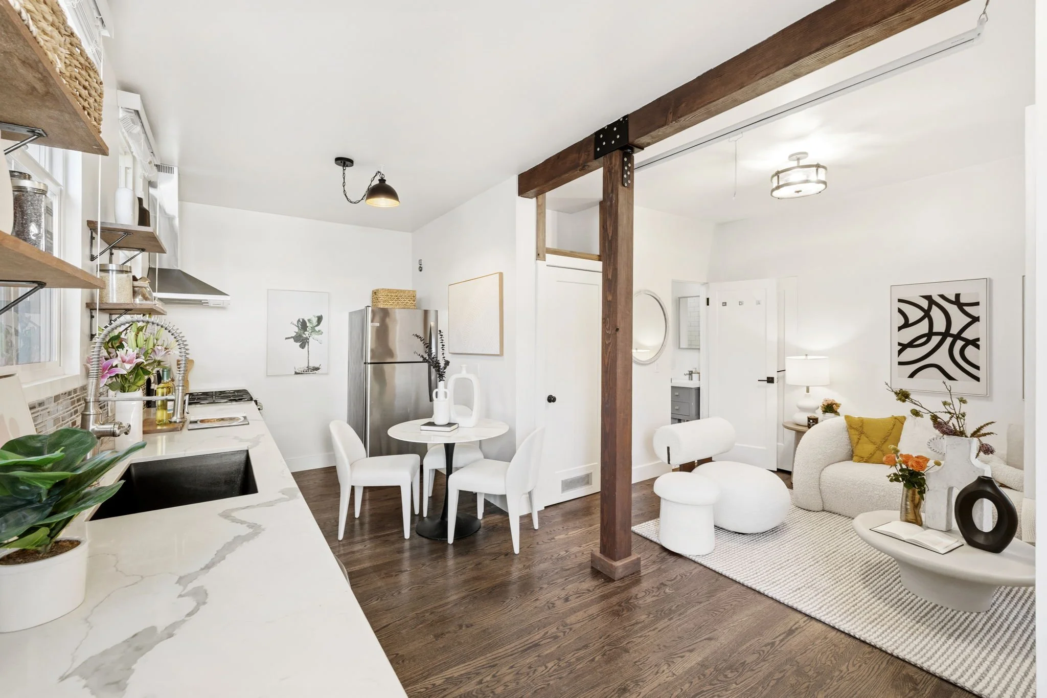Open-concept living room and kitchen with white walls, wooden accents, and modern furniture. Kitchen has open shelving, a marble countertop, and a stainless steel refrigerator. Living area features a white sofa, a round coffee table with vases of flowers, and wall art. Wood flooring and natural light create a bright, minimalist aesthetic.