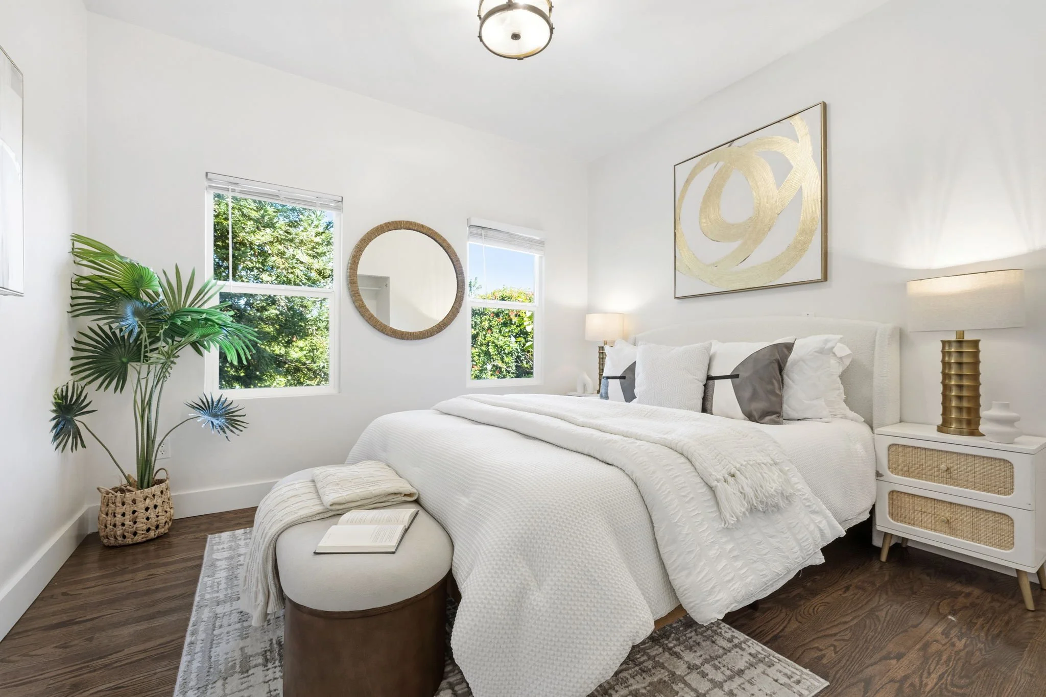 Bright, modern bedroom with white bedding and decor, a large rectangular white headboard, two bedside tables with lamps, a decorative mirror on the wall, a large abstract gold artwork, a potted plant, a beige ottoman with an open book and blanket, two windows showing greenery outside, and hardwood floors with a patterned rug.
