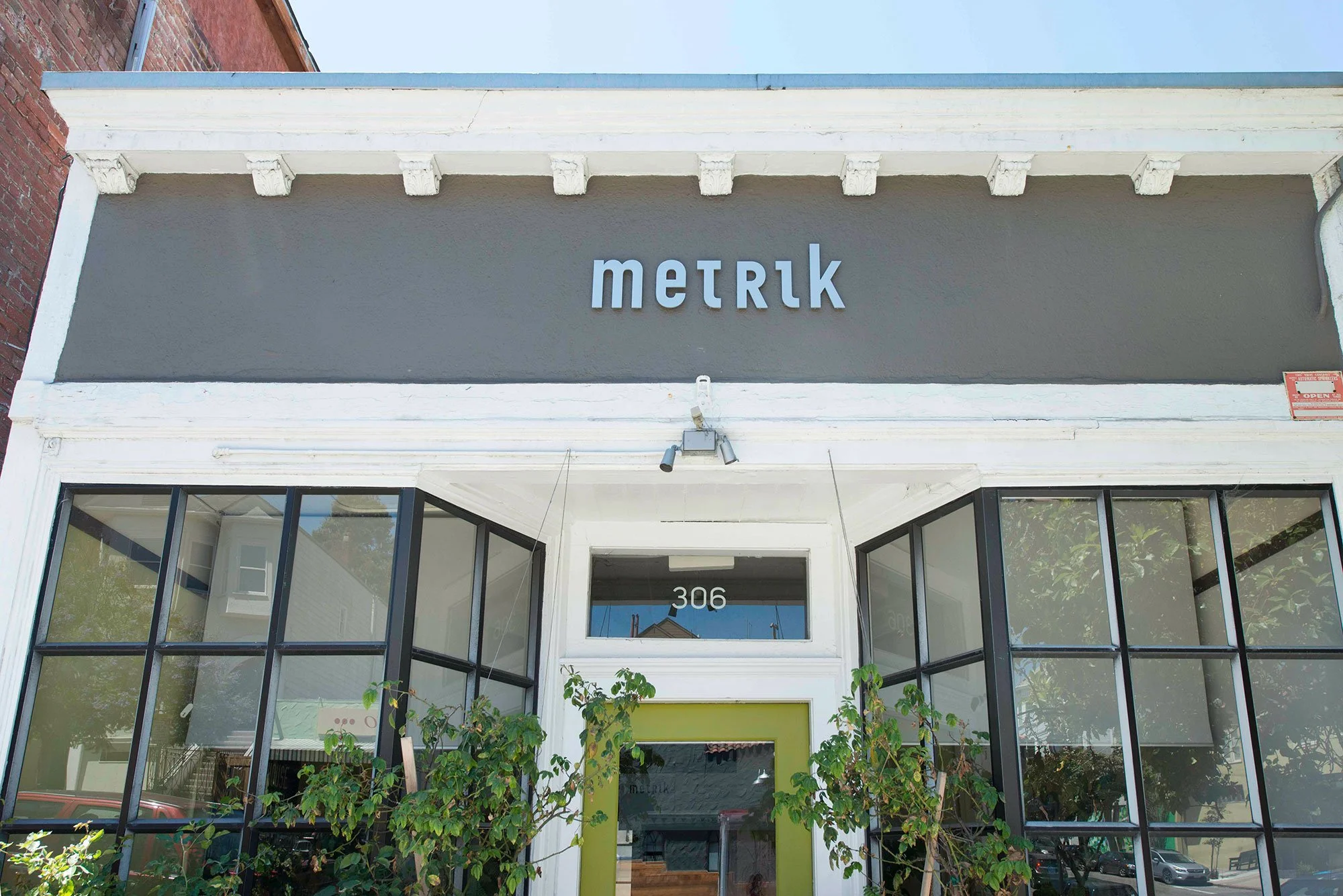 Exterior view of a storefront with the sign 'metrik' above the entrance, which is framed by large glass windows and green plants.